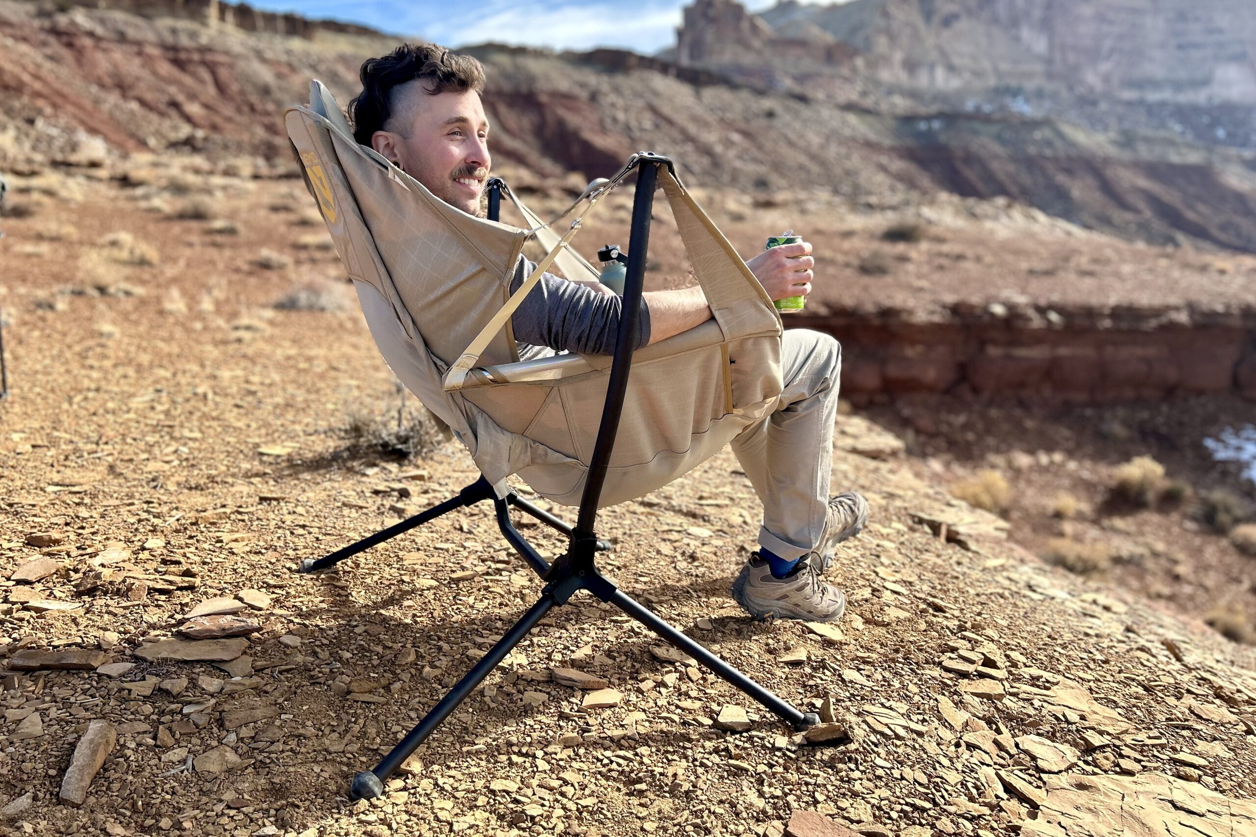 A man sits in a camping chair having a canned beverage in a sunny desert setting.