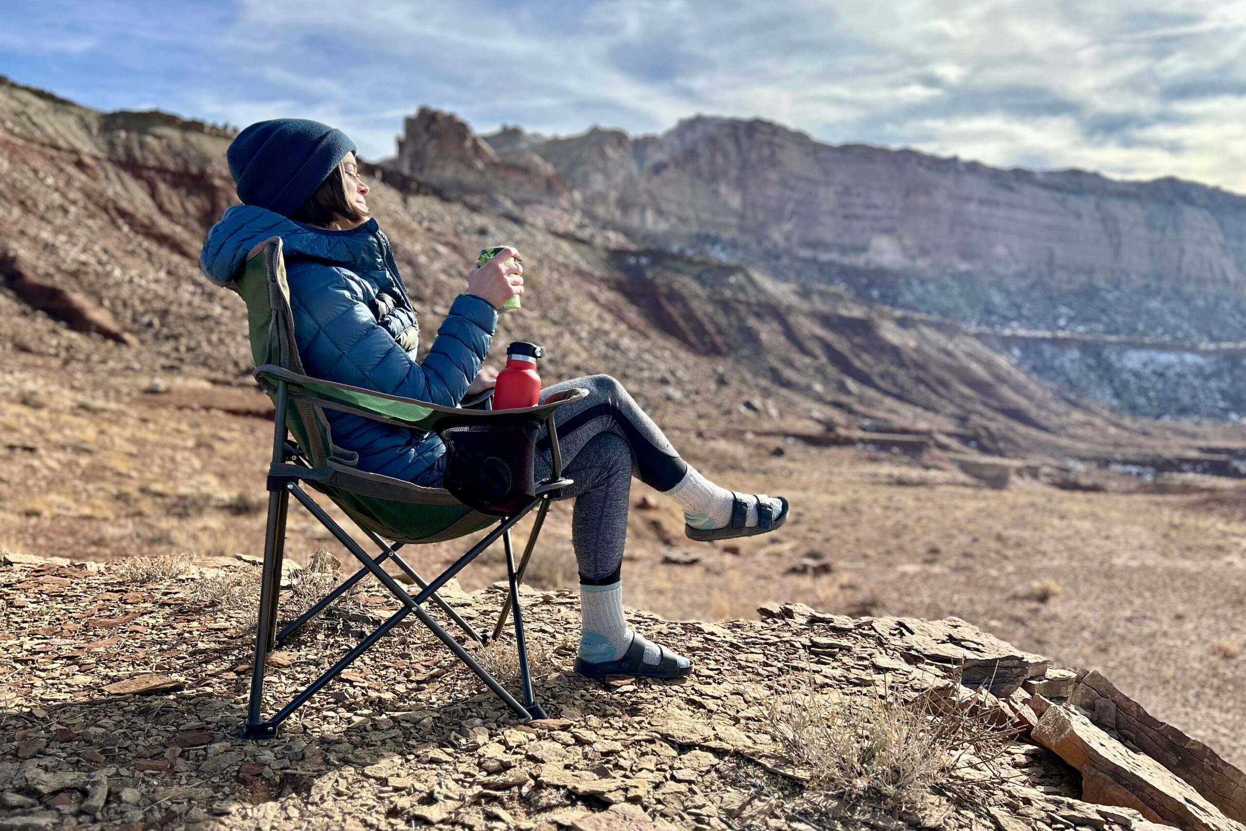 A woman sits in a camping chair in the desert drinking from a can.