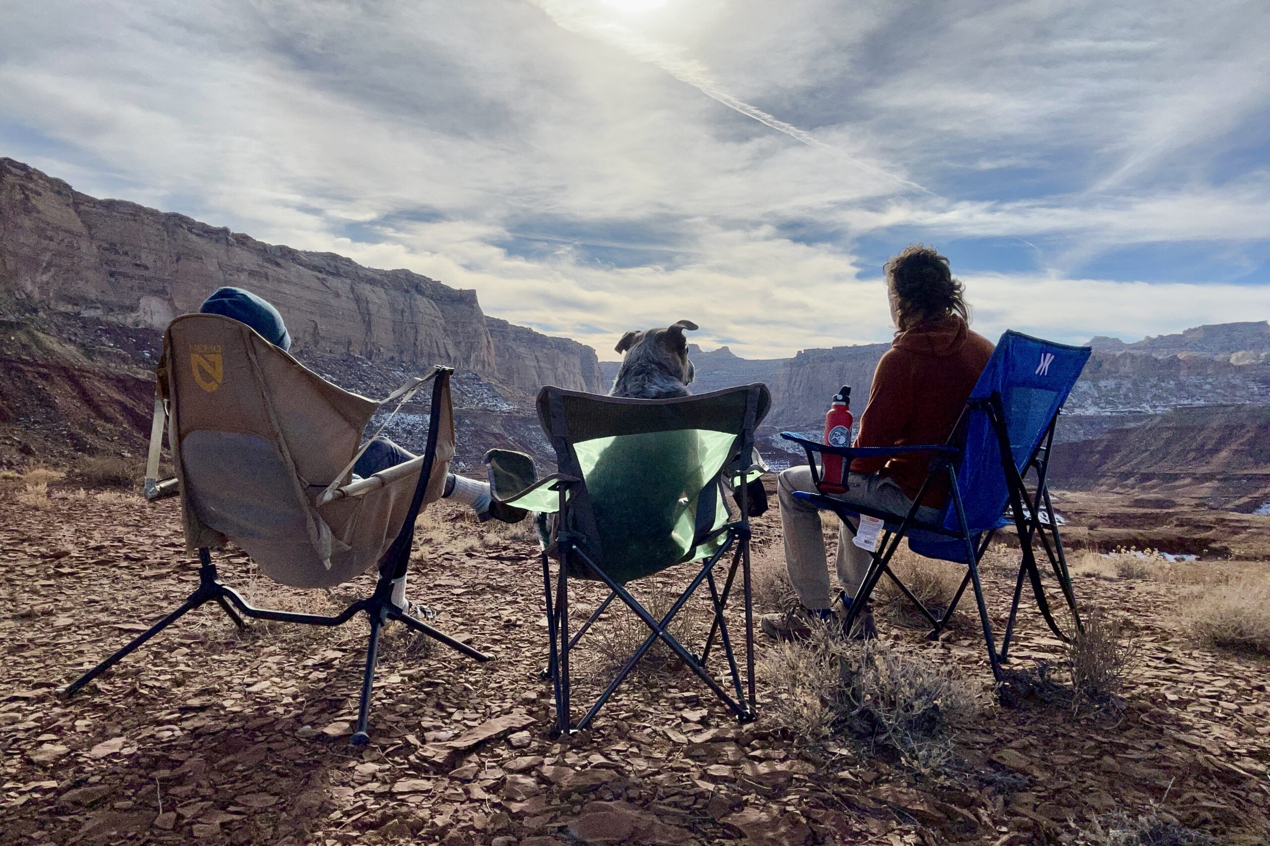 Two people and a dog sit in three camping chairs in a sunny desert setting.