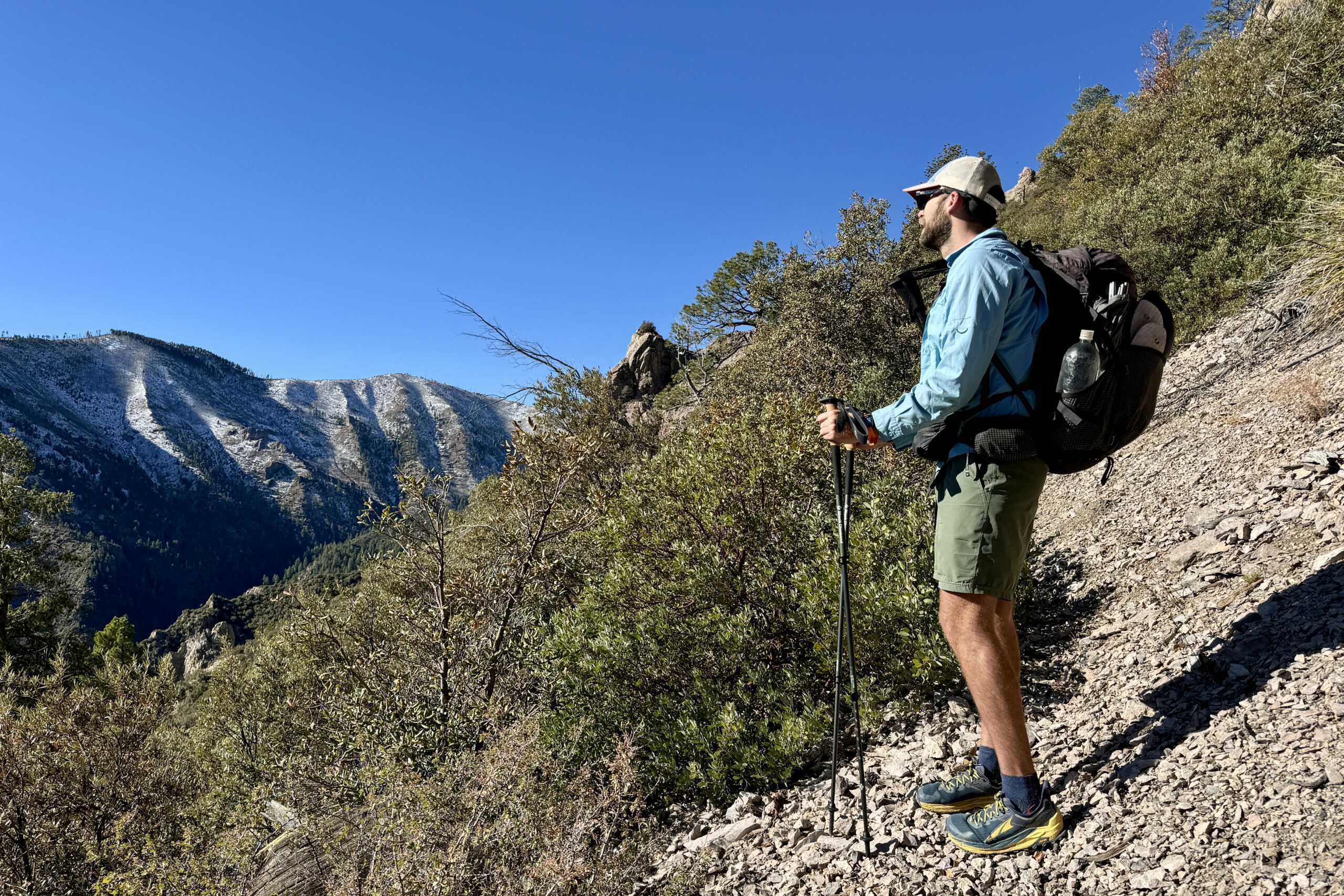 A person wearing a backpack and holding trekking poles is standing on a trail looking at mountain views.