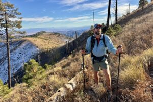 A person wearing a backpack and holding some poles is standing on a trail catching their breath with mountain views and a desert behind them.