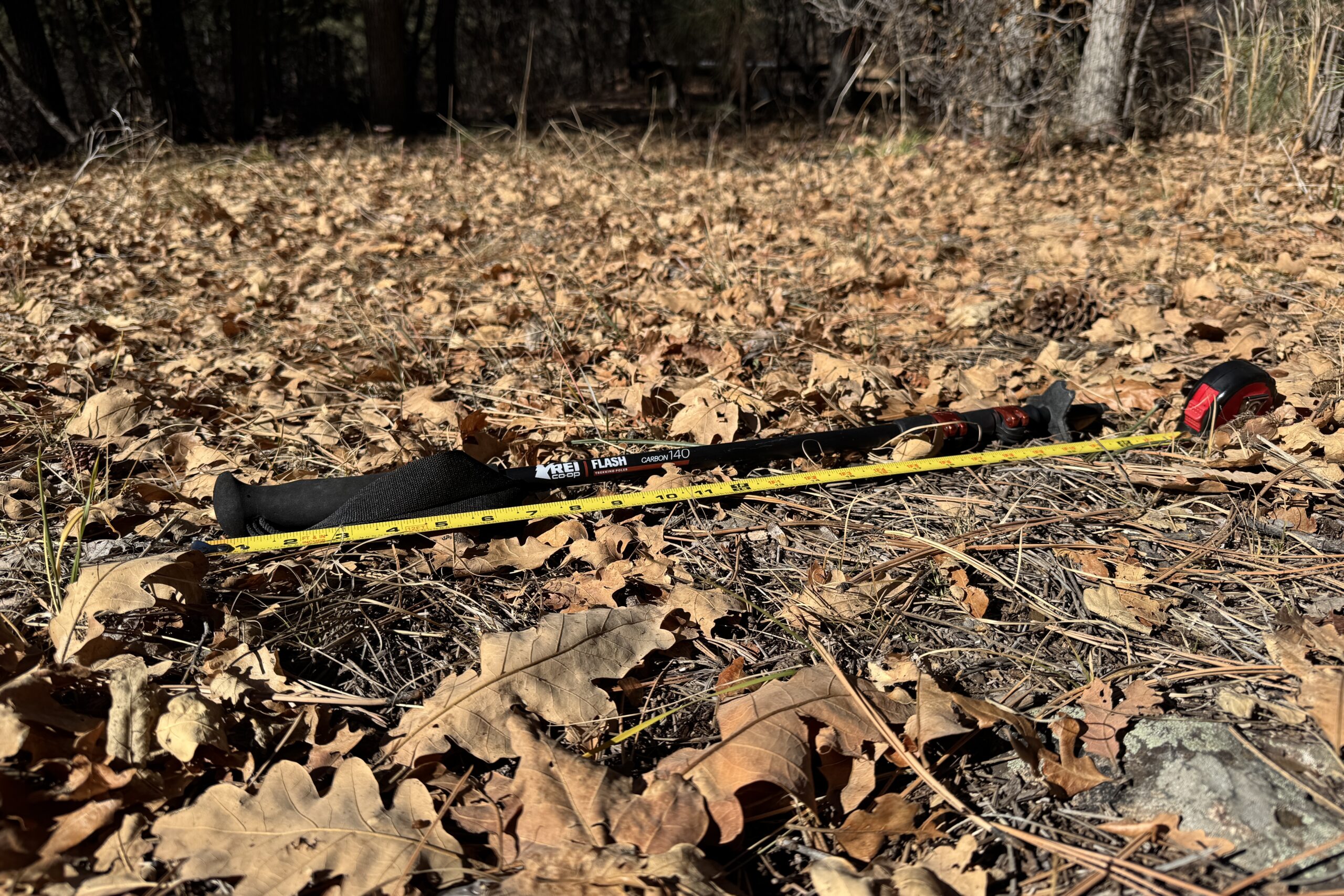 The REI Flash Carbon pole is laying on the ground in some leaves with a measuring tape next to it.