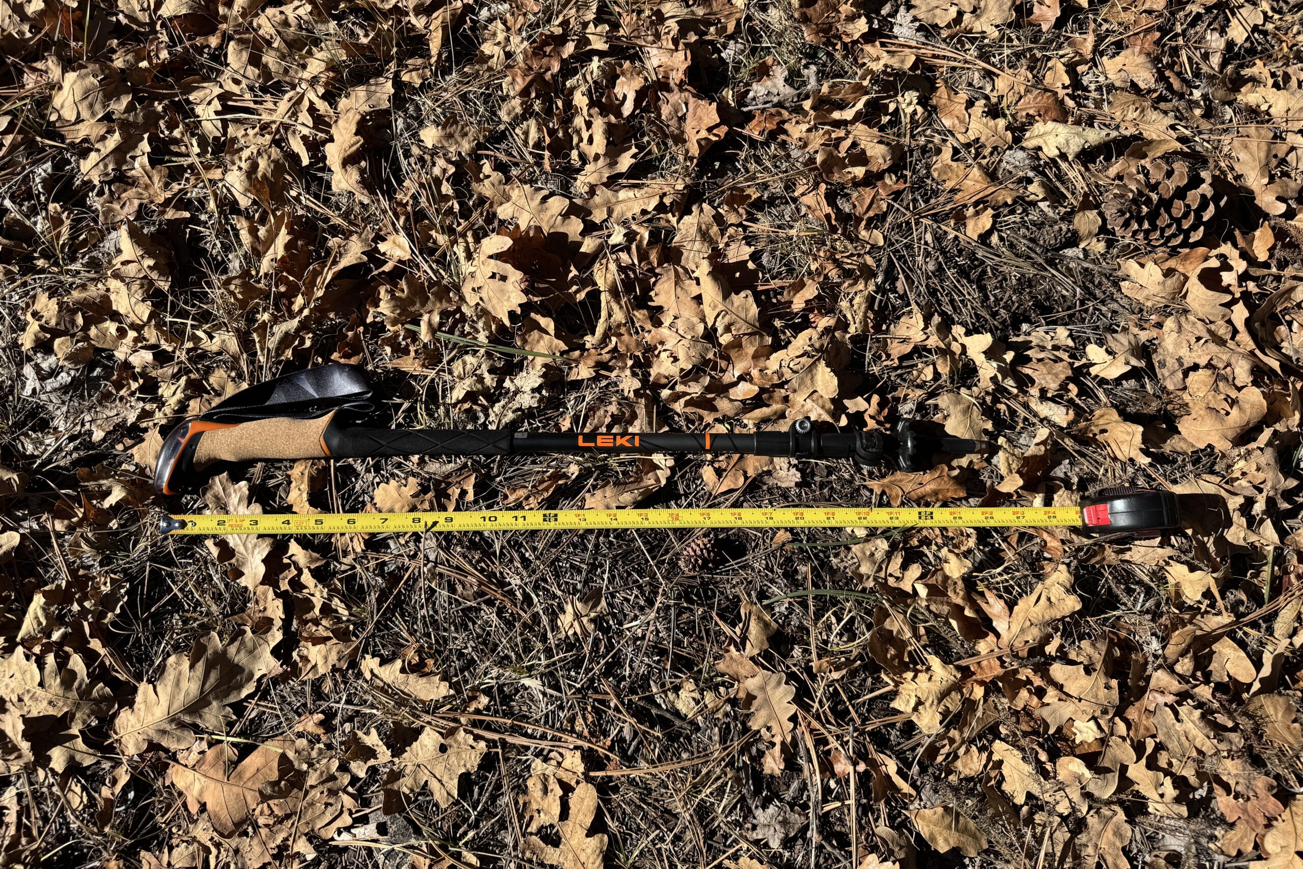The Leki Makalu Cork Lite pole is laying on the ground in some leaves with a measuring tape next to it.