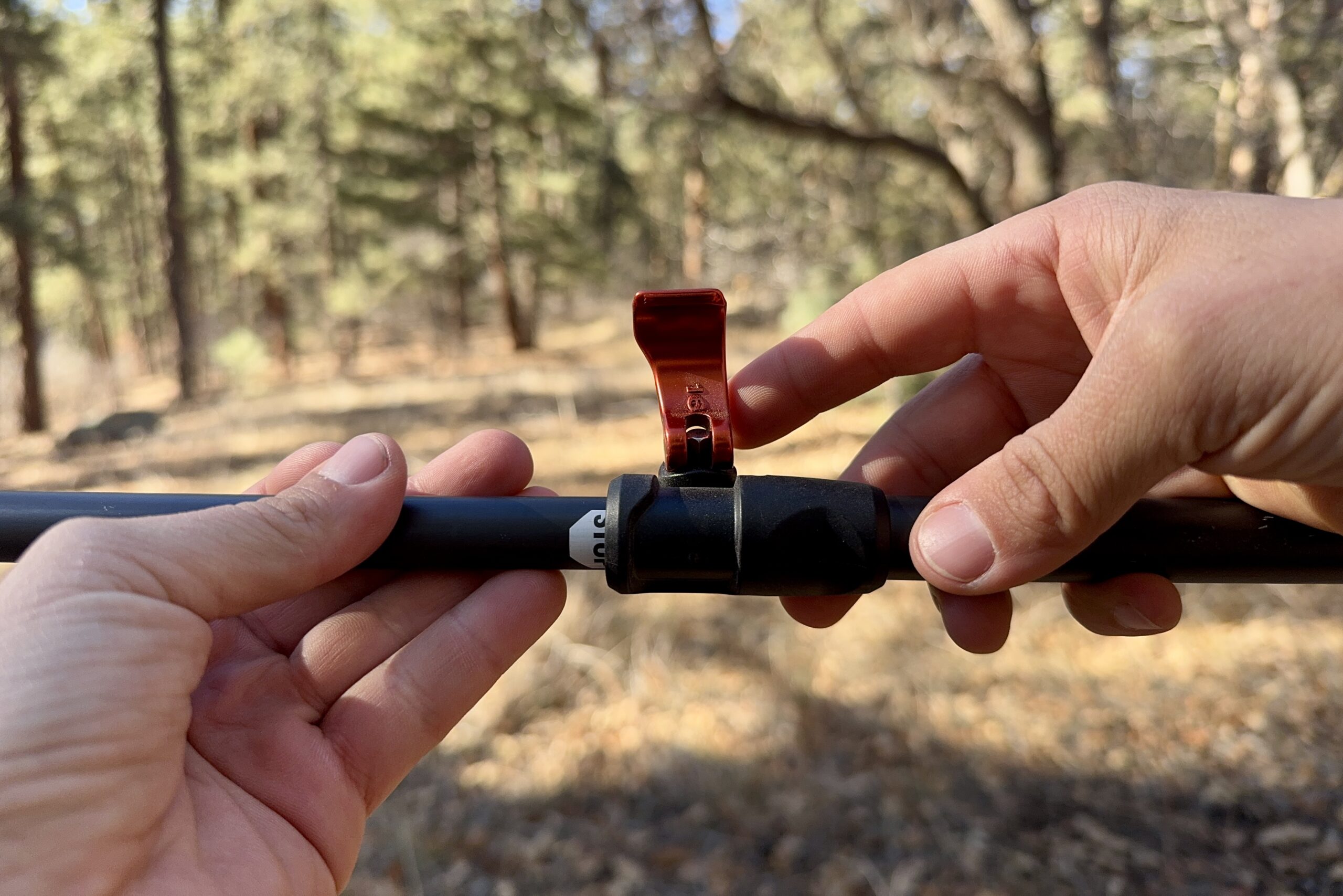 A close up of two hands that are closing a flick lock on a trekking pole. There is a forest in the backround.