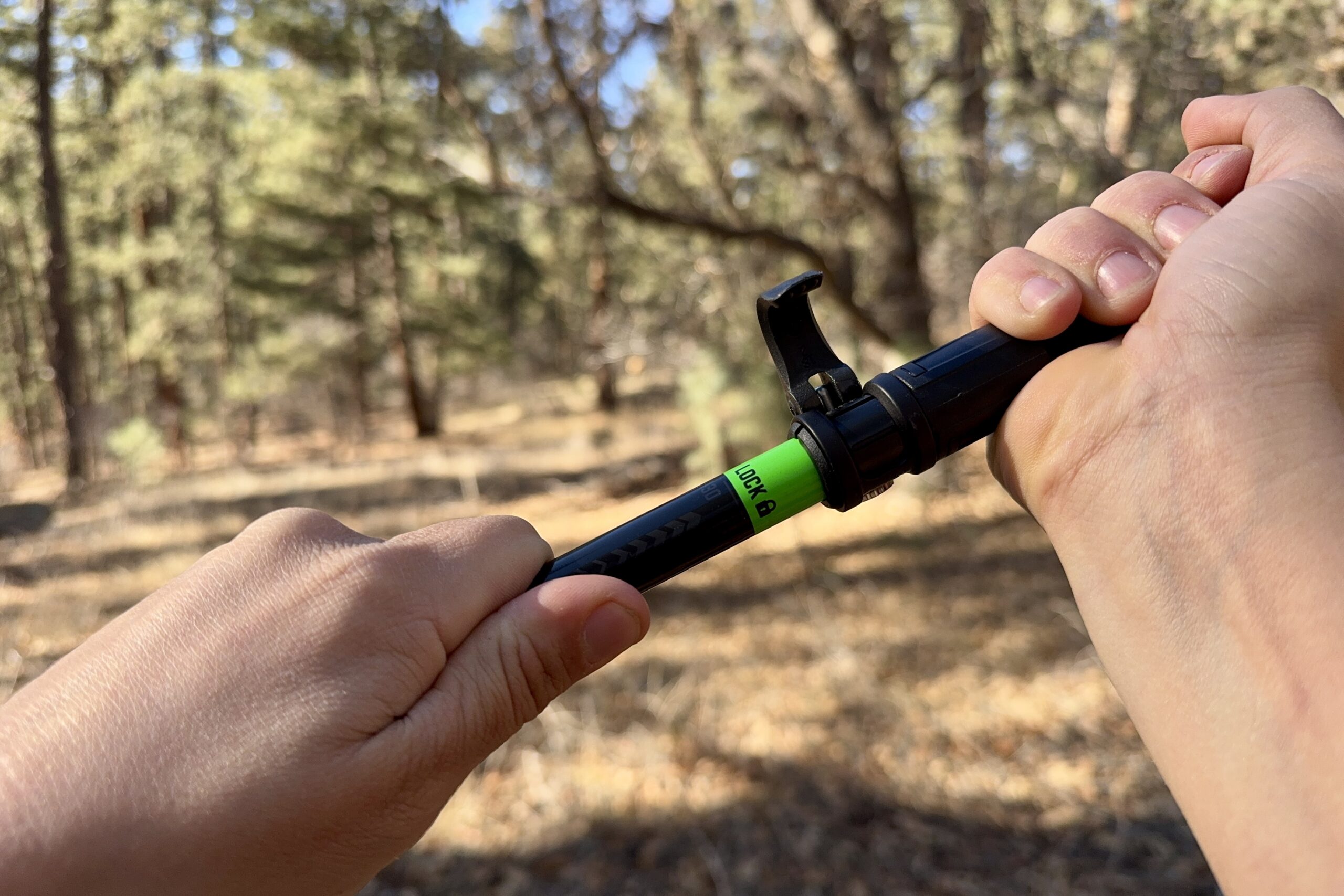 A close up of two hands pulling on a trekking pole to engage the tension lock on a pair of folding style poles. There is a forest in the background.