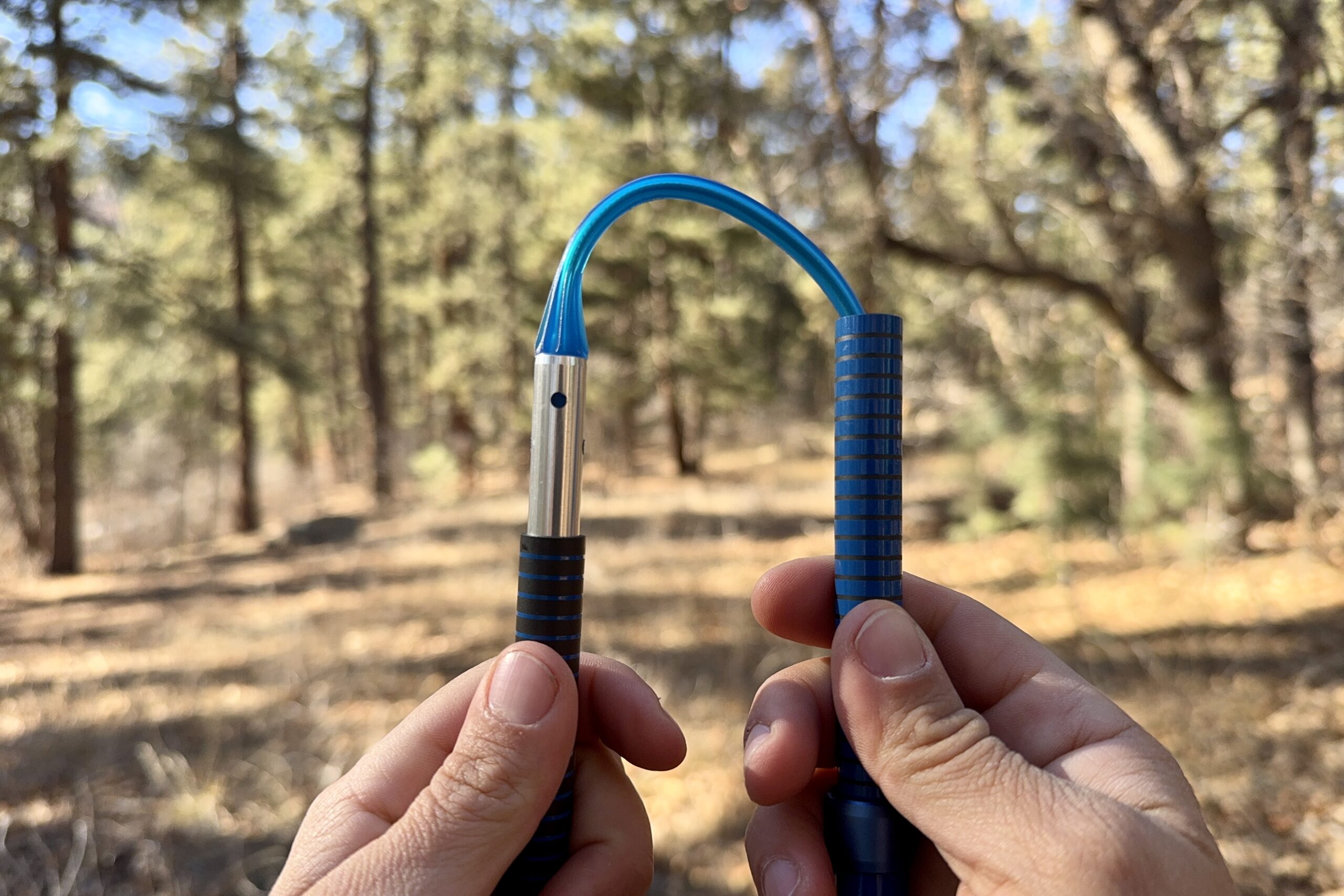A close up of two hands holding a folding trekking pole and showing the inside cable. There is a forest in the background.