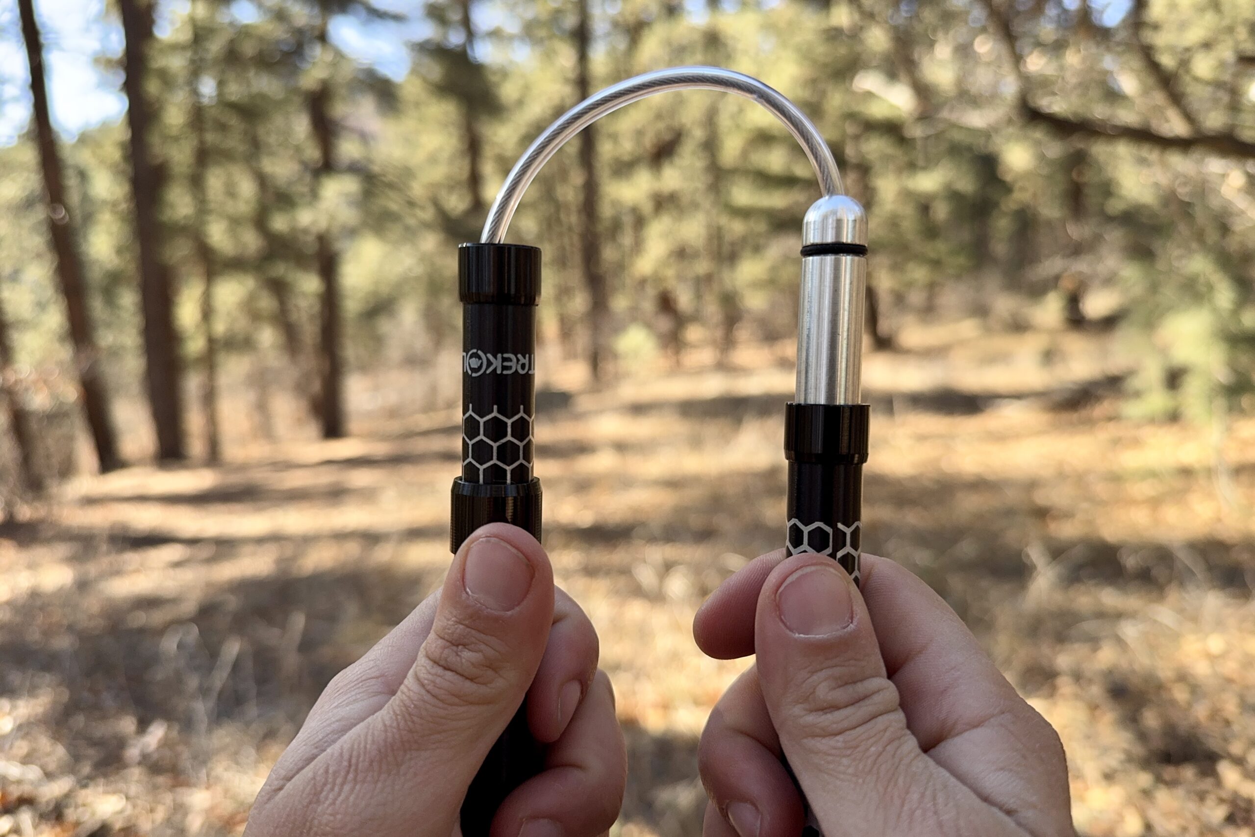 A close up of two hands each holding a shaft of a folding style pole. There is a cable cord connecting the shafts and a forest in the background.
