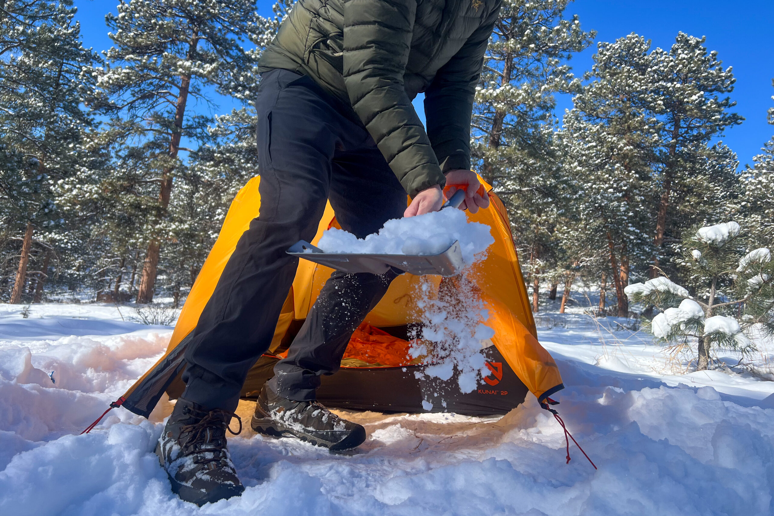 A close-up of a camper shoveling snow near the orange Nemo Kunai 2 tent, showcasing the use of a lightweight aluminum snow shovel.