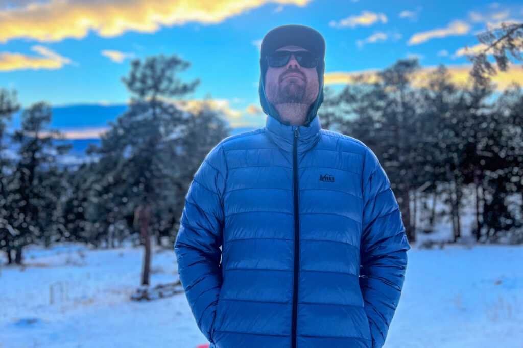 A man stands facing the camera wearing a blue REI 650 Down Jacket, zipped up with a hood underneath a cap, set against a snowy winter forest at sunset.
