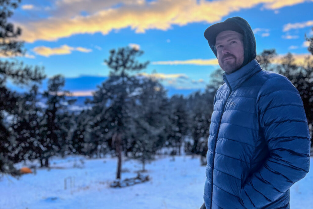 A man in a blue REI 650 Down Jacket stands with his hands in his pockets, gazing off into the snowy forest during an early winter evening.