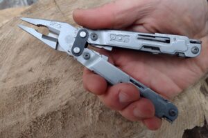 The silver SOG PowerAccess in hand, held above a log, with its needle nose pliers extended.