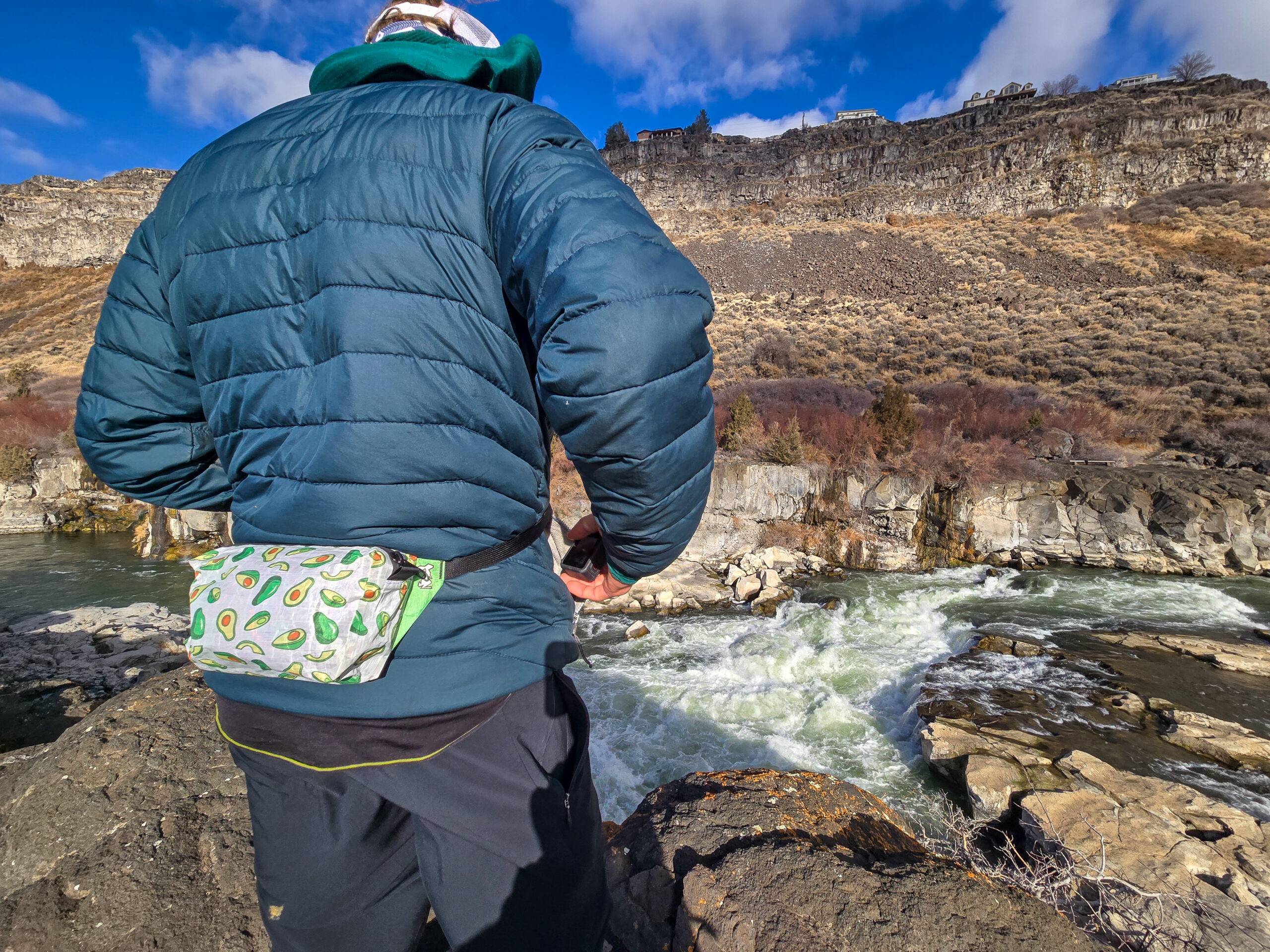 man overlooking a whitewater river rushing through a canyon on a sunny day while wearing a high tail designs fanny pack over a down jacket