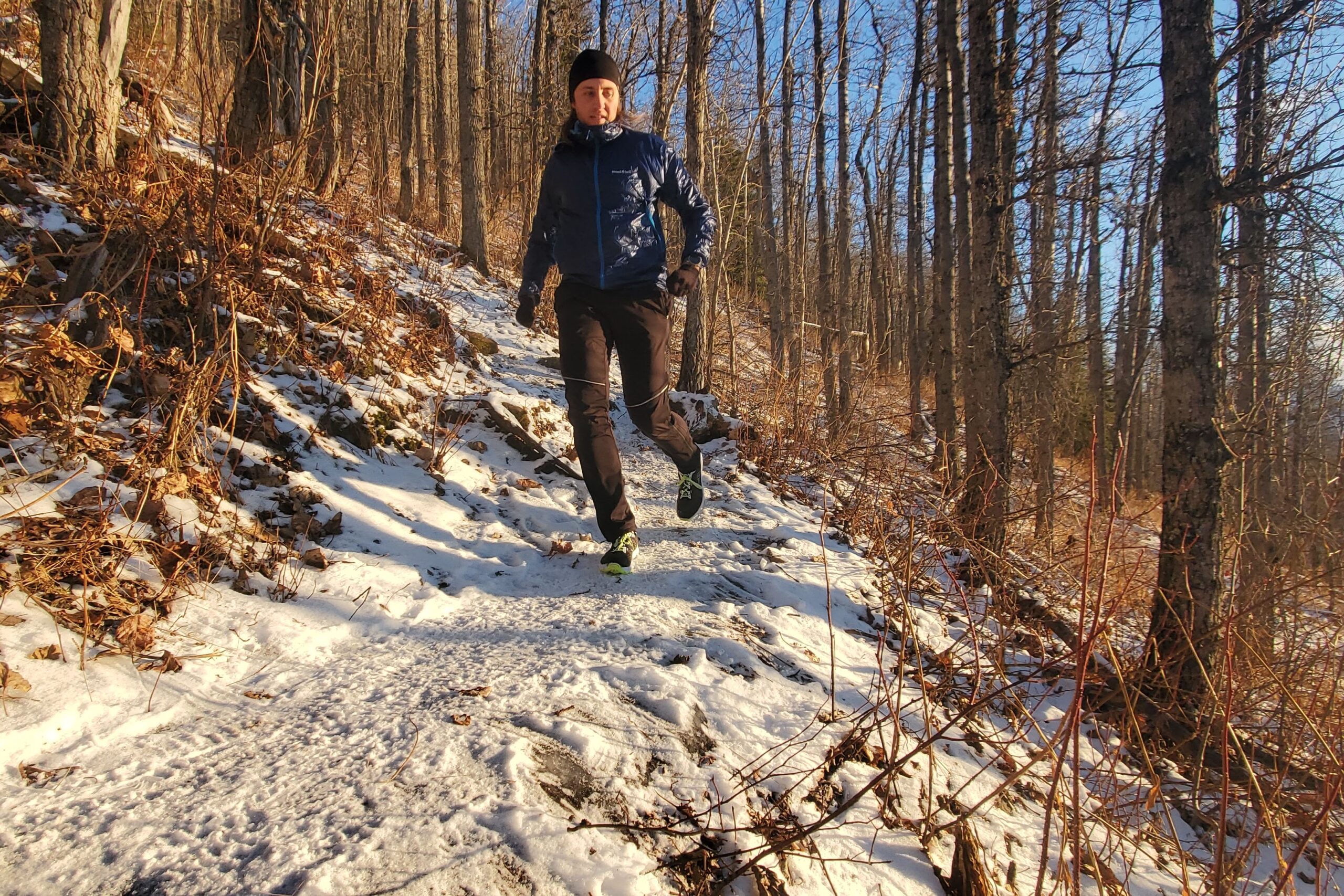 A man runs down a snowy trail in the sunshine wearing a blue jacket.