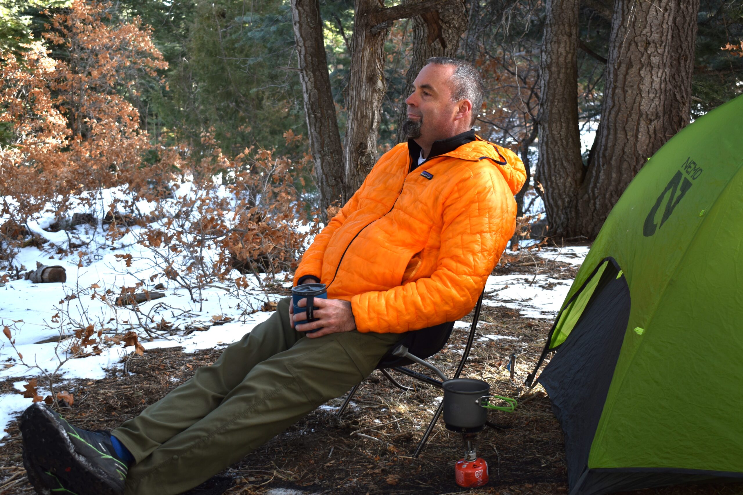 A backpacker holds a coffee cup while sitting on a backpacking chair next to a tent with snow on the ground