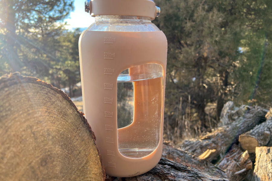 A large beige BLUEGO 84-ounce glass water bottle with measurement lines, sitting outdoors on a log.