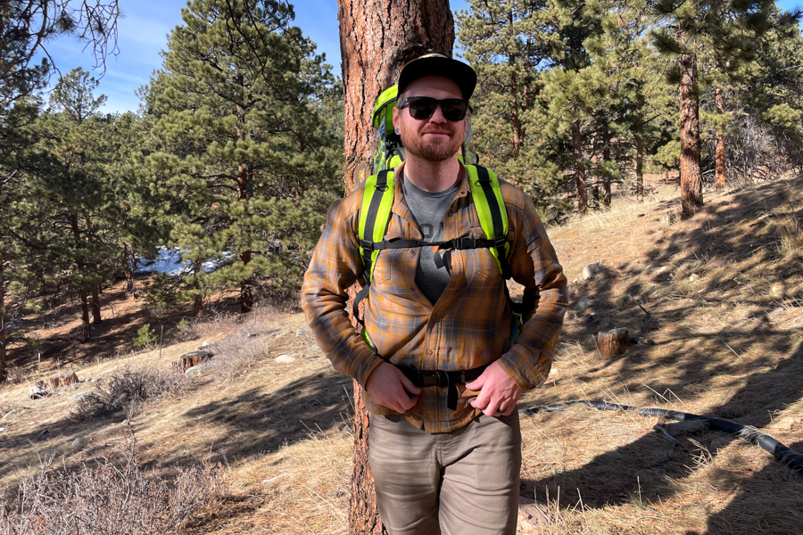 A man standing outdoors wearing a bright green child carrier, smiling while leaning against a tree in a forested area.