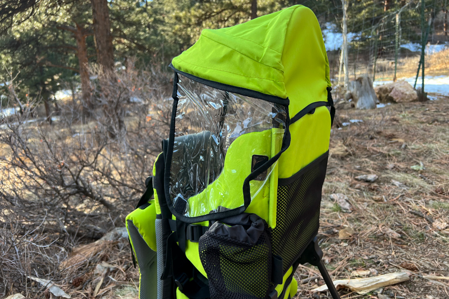 Side view of a bright green child carrier with a transparent rain cover attached, displayed outdoors in a forest setting.