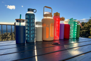 A variety of reusable water bottles arranged on a table with a scenic mountain sunset in the background, showcasing different shapes, colors, and designs.