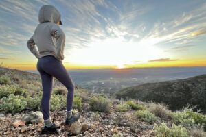 A woman stands before the setting sun with her hands on her hips.