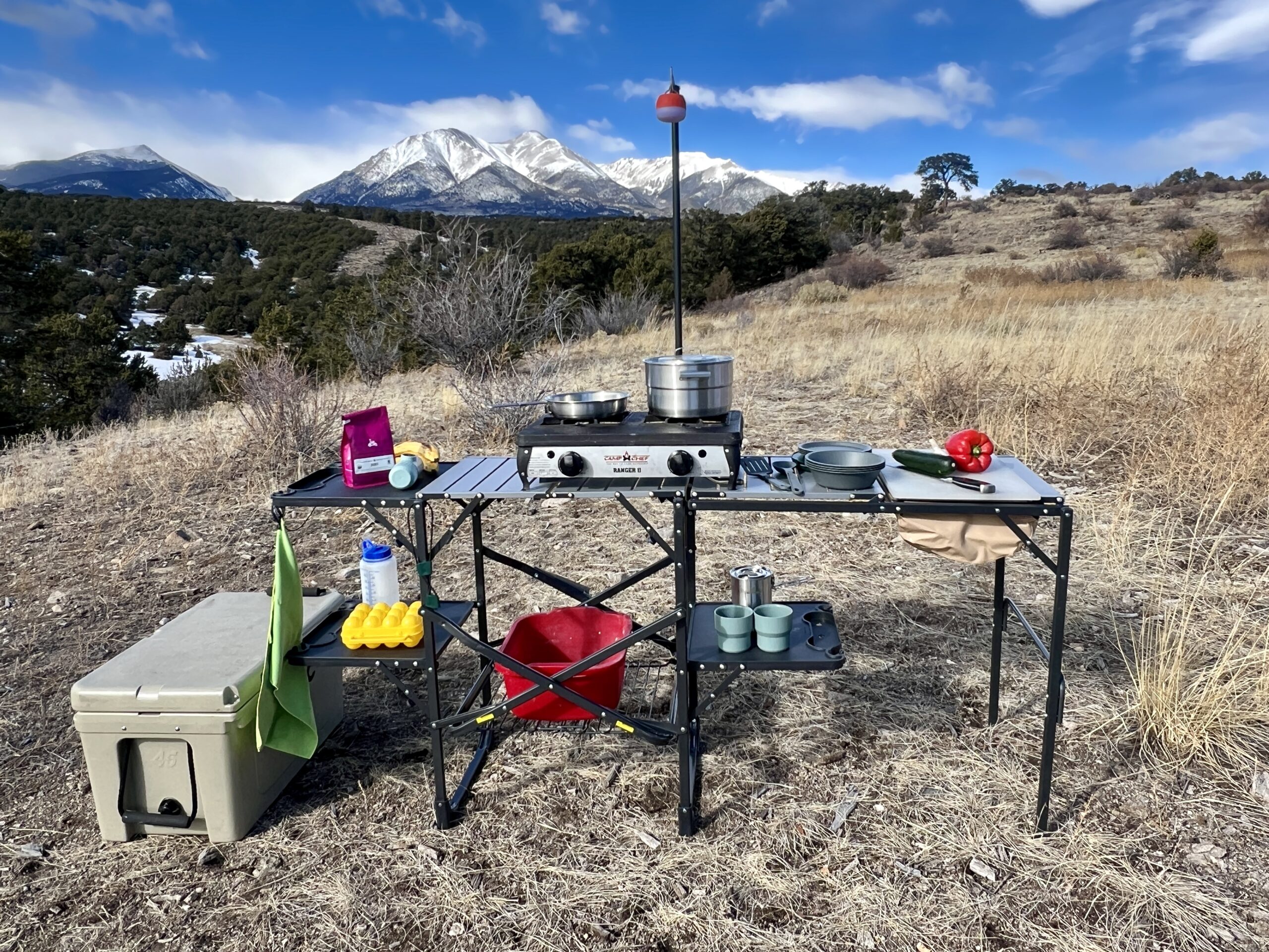The GCI Master Cook Station stands outside with a mountain backdrop. Its shelves hold kitchen items and an outdoor stove. A cooler is on the ground next to it.