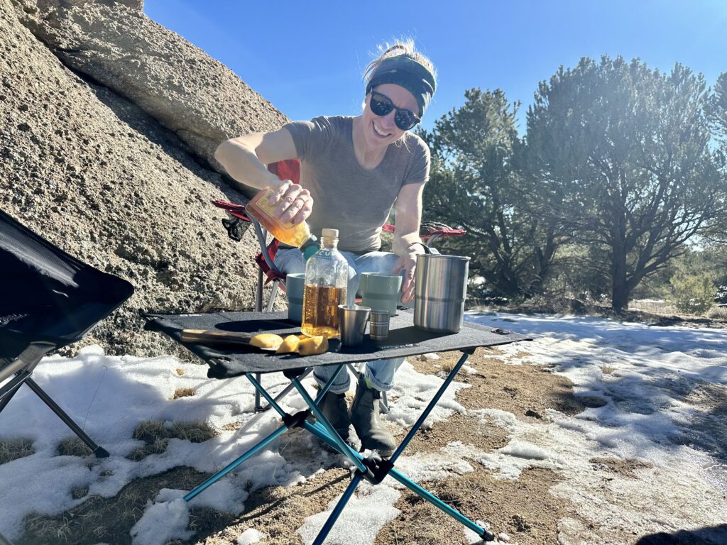The Helinox Table One Hard Top has beverages and sliced lemons on top of it. A person is smiling and squeezing a bottle of honey. They are wearing sunglasses, a bandana, and a brown t-shirt. The backdrop has trees and a large boulder and there is snow on the ground.
