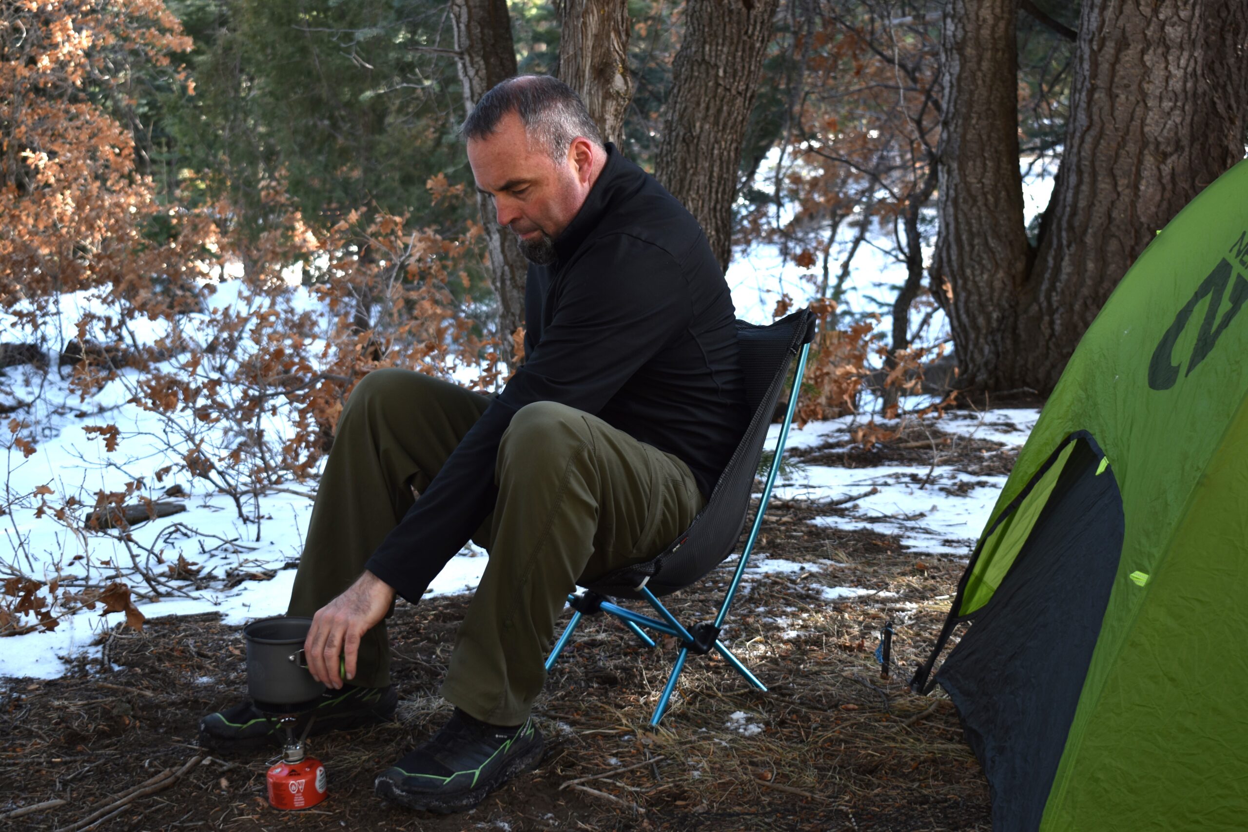 A male backpacker leans forward on the Helinox Chair Zero to reach a pot on his camp stove with snow on the ground