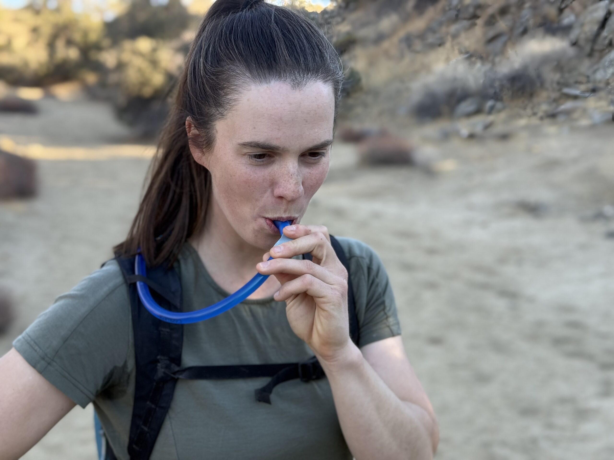 Hiker drinks from a hydration bladder standing in a sandy wash.