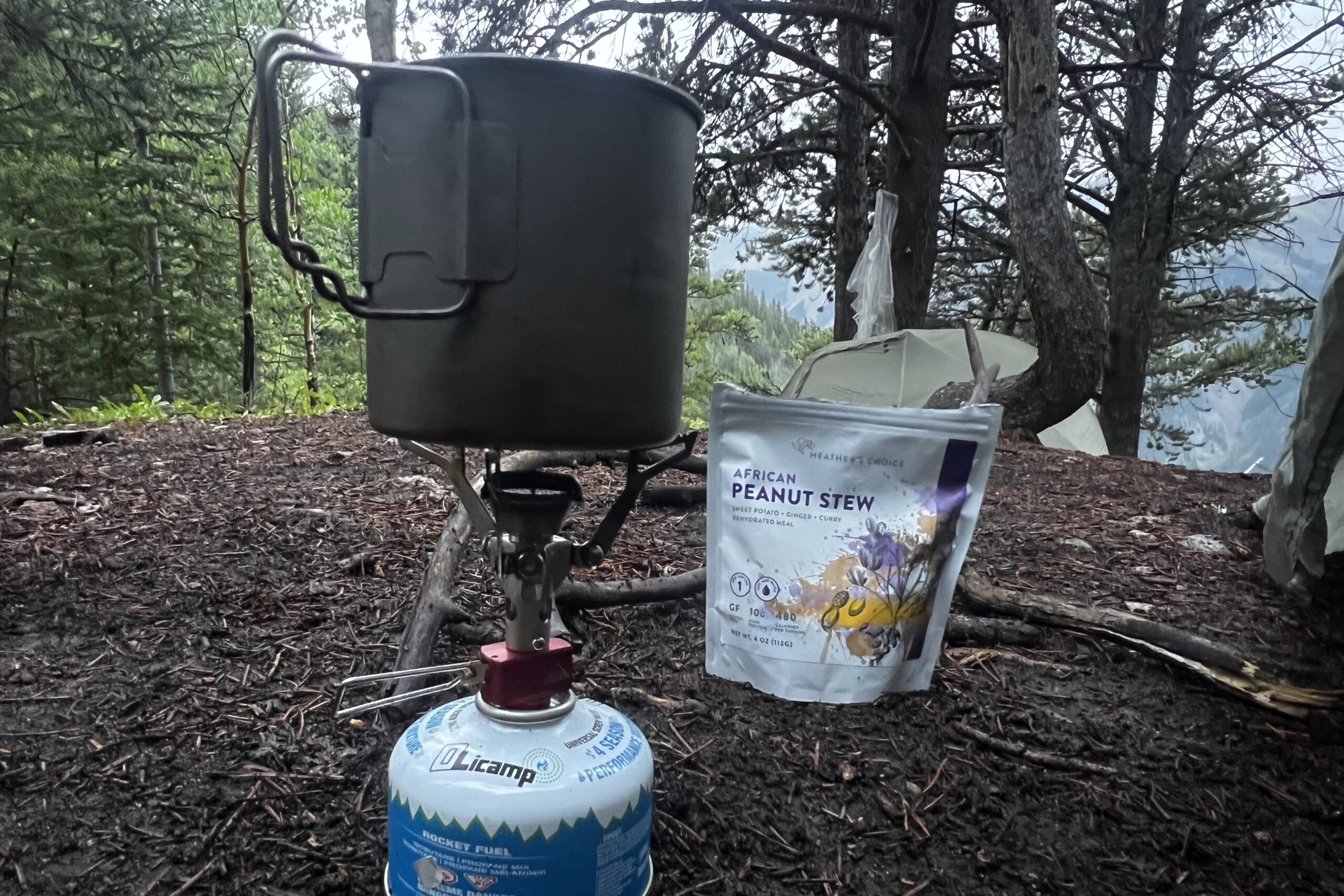 A backpacking pot sits on a stove with a dehydrated meal nearby and trees in the background.