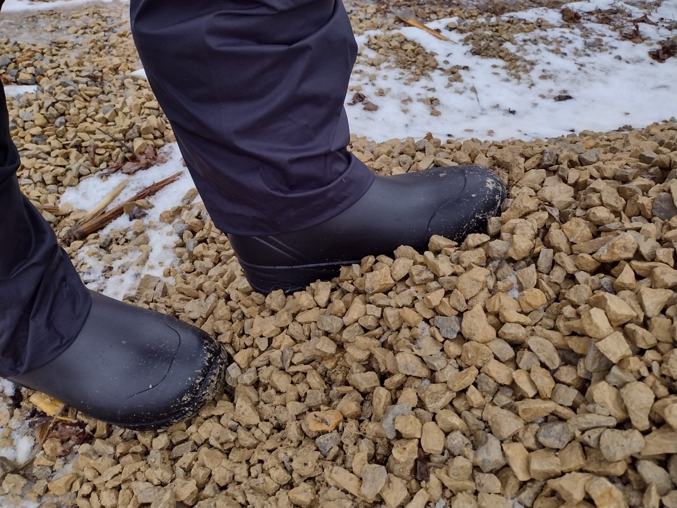 Ground level view of someone wearing the black Bozeman Mid boots walking on a gravel pile.