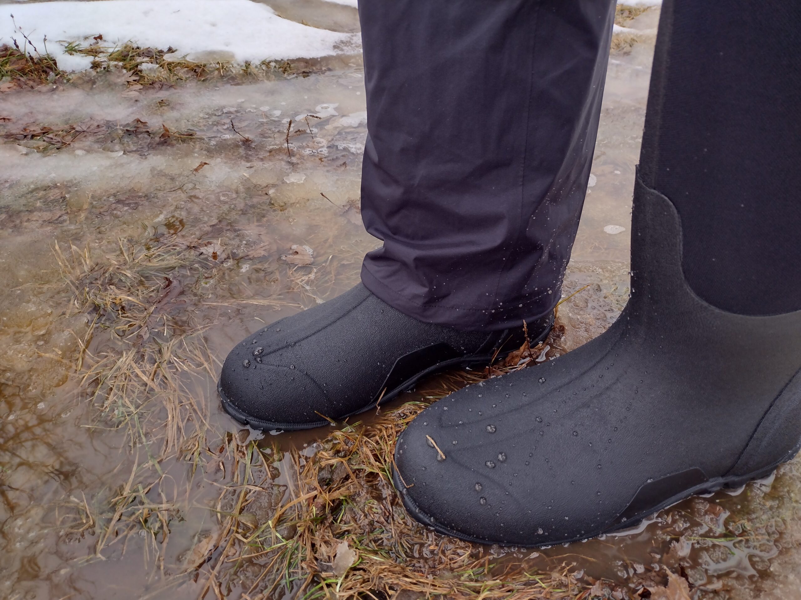 Ground level view of someone wearing the black Bogs Classic High standing on soggy ground.