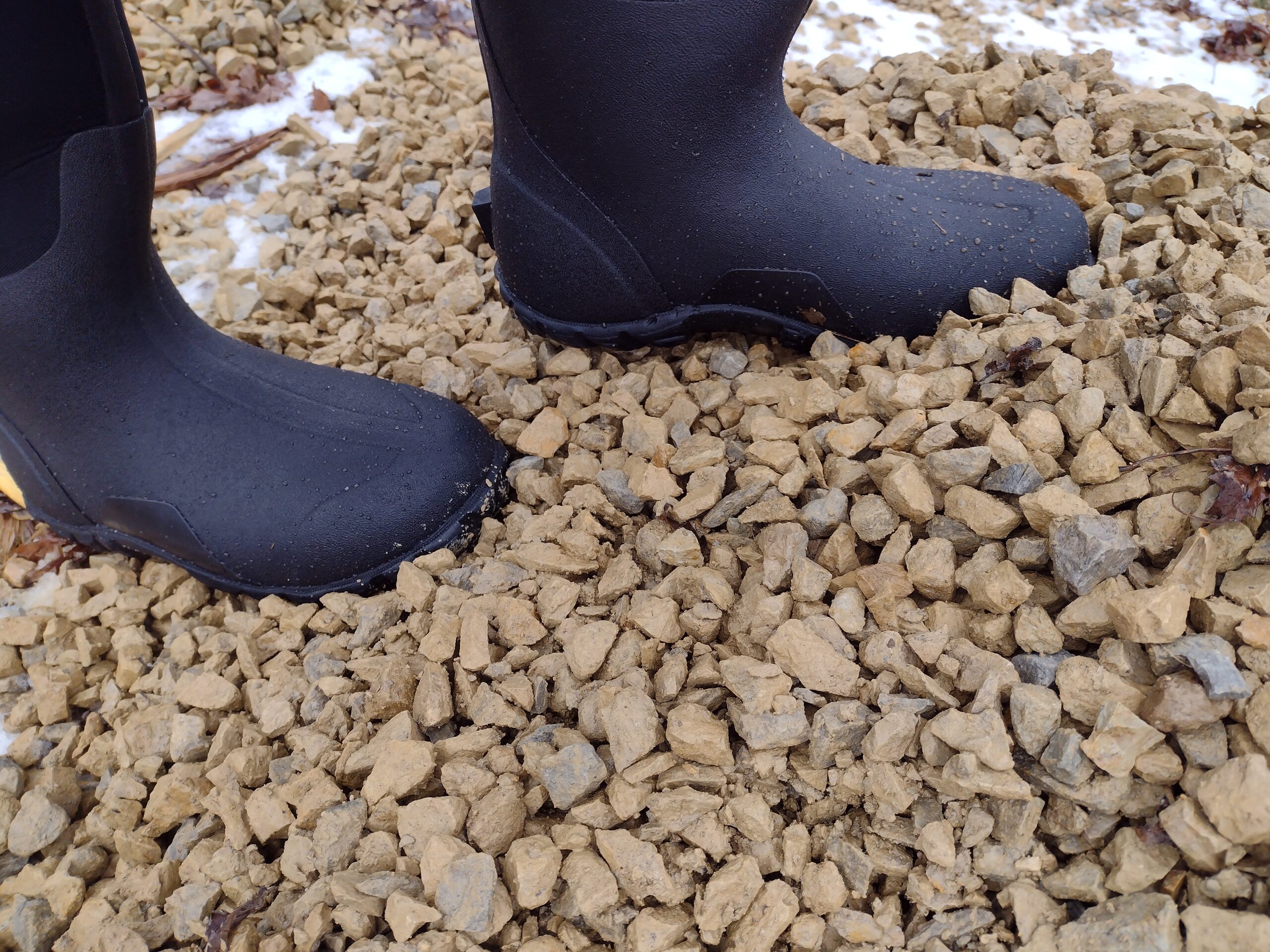 Ground level view of someone wearing the black Bogs Classic High standing on a gravel pile.