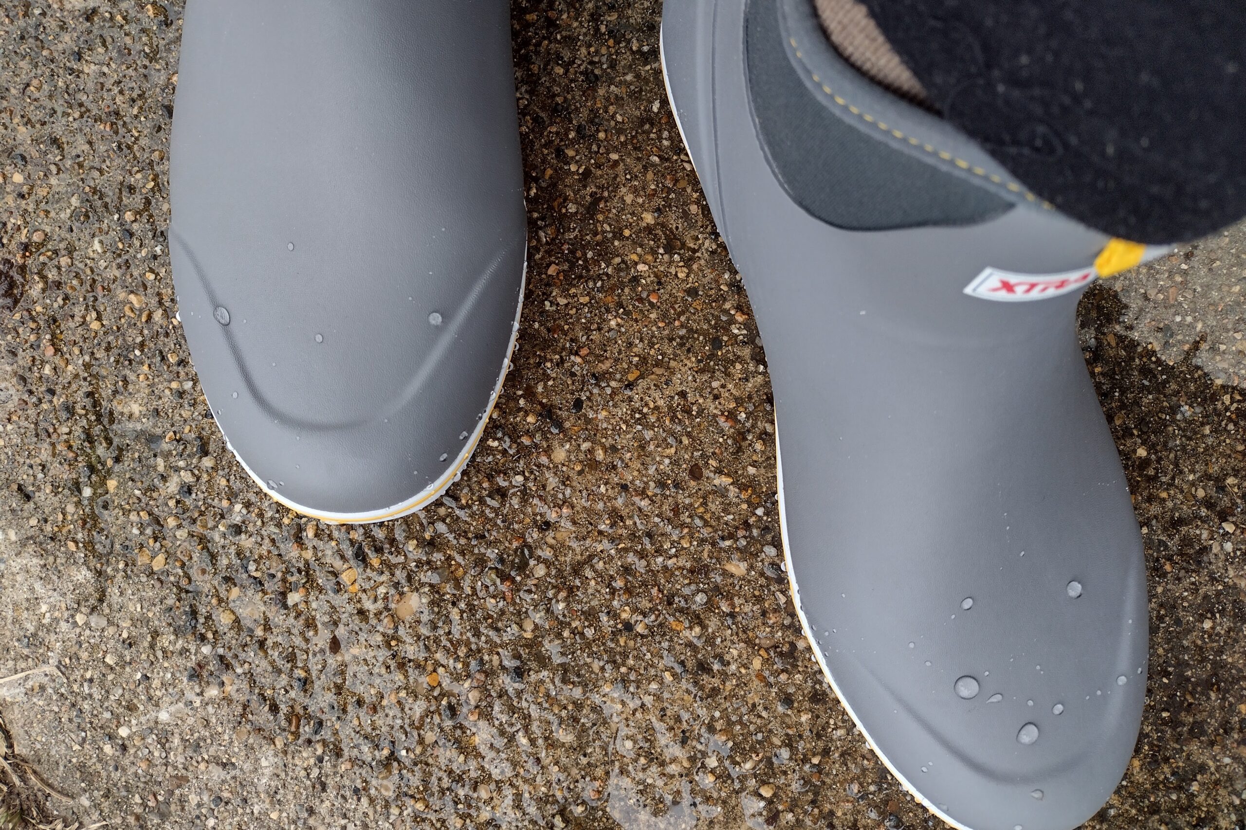 Ground level view of someone wearing the gray XTRATUF Ankle Deck Boot on a wet sidewalk.