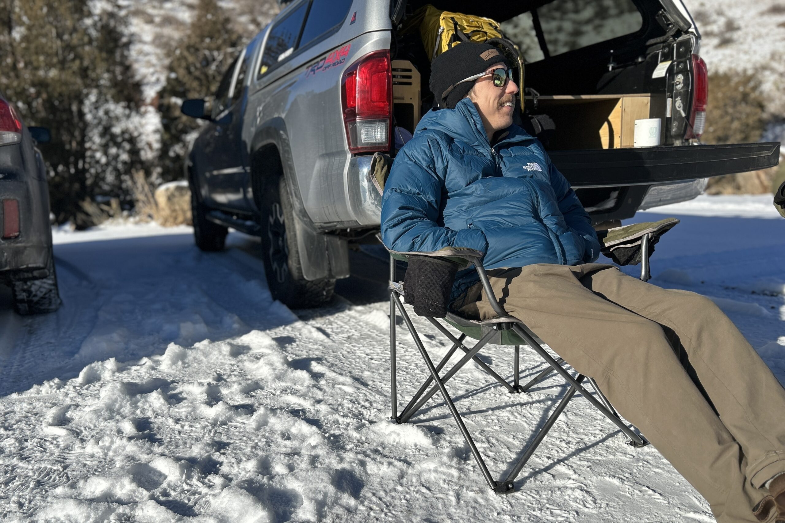 A man sits in a camping chair in a snowy parking lot while smiling.