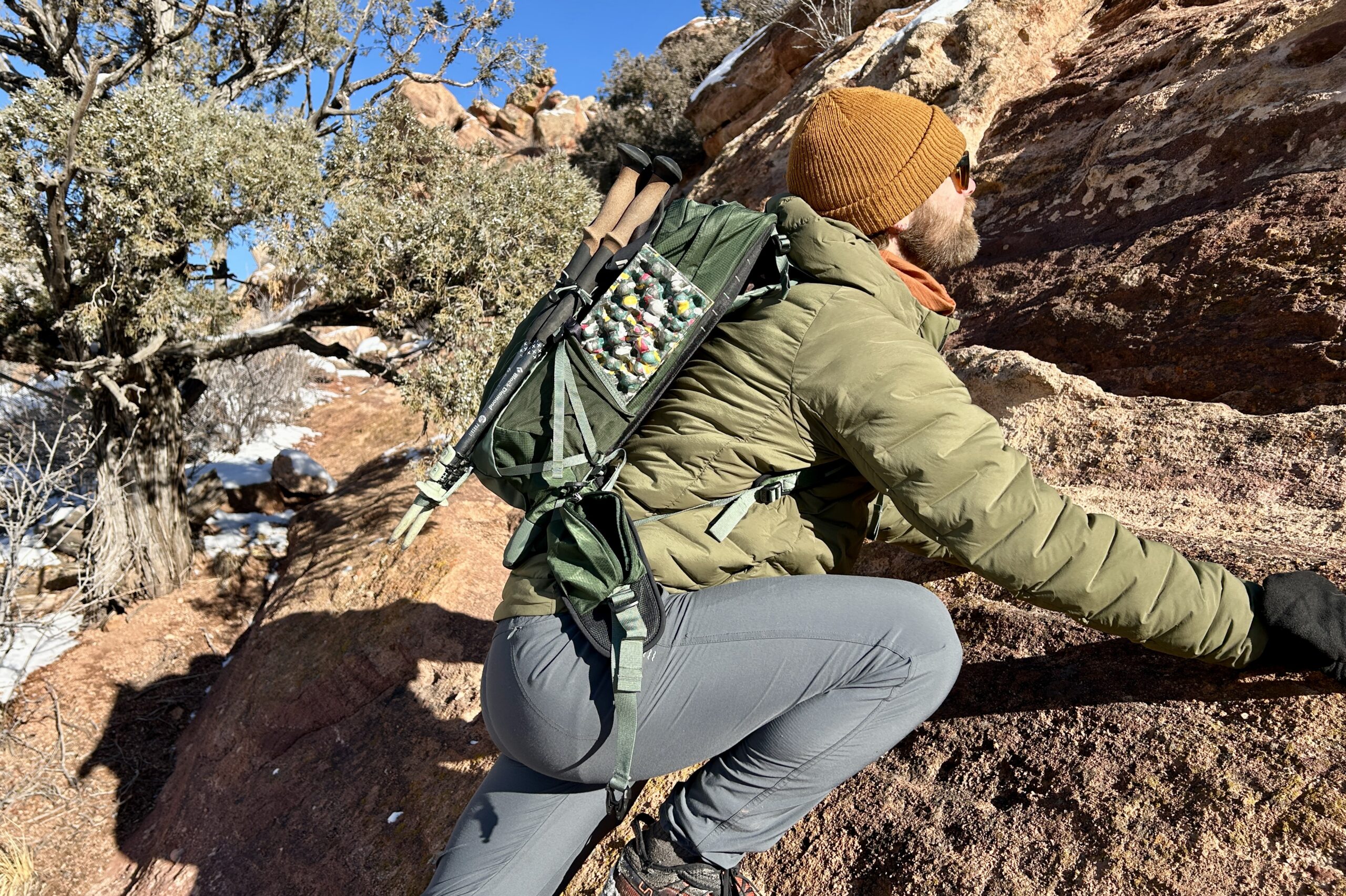 A man scrambles up a rock wearing a jacket, hat, and a backpack with hiking poles attached to it.