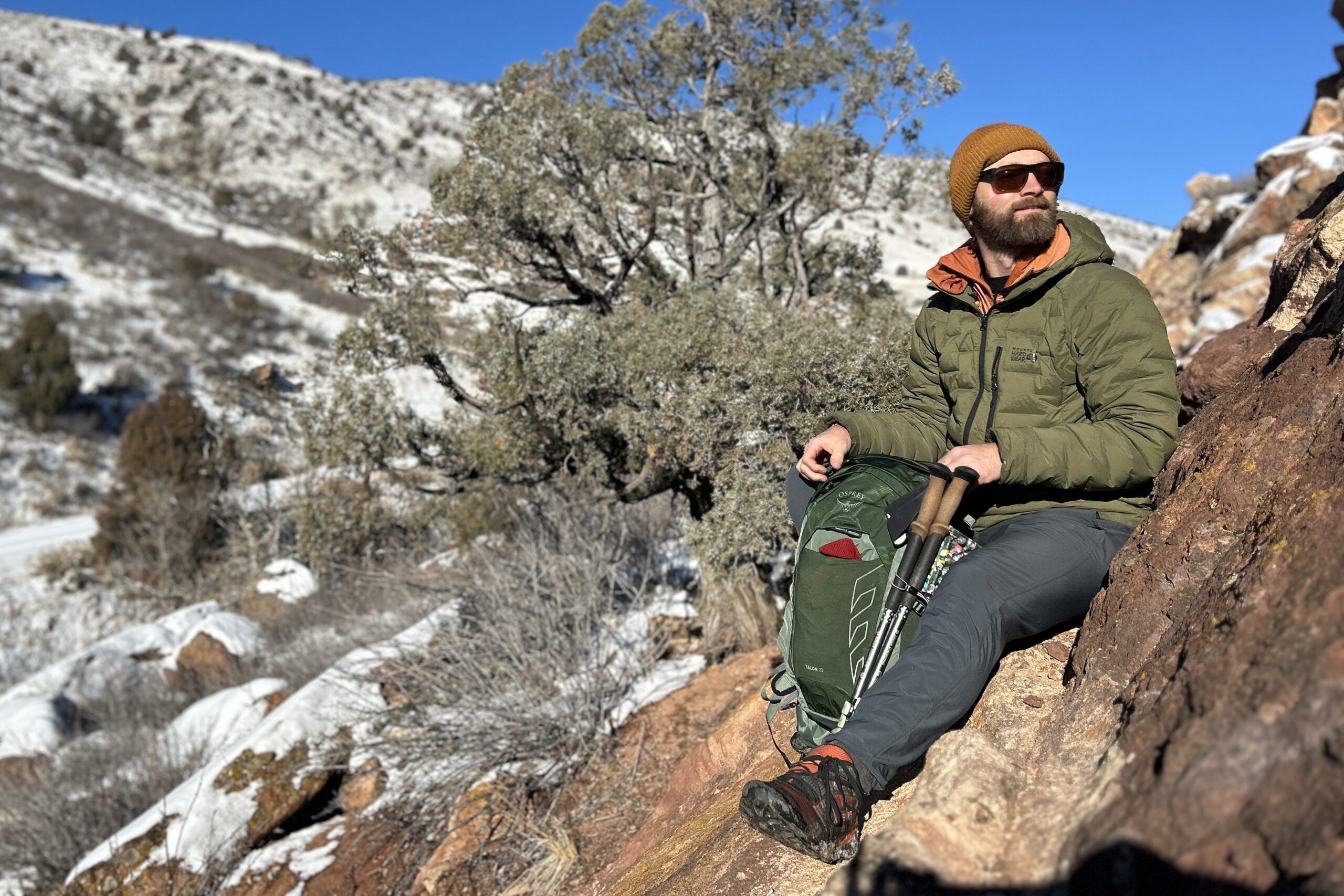 A man sits on red rocks in a snowy setting with his backpack between his legs.