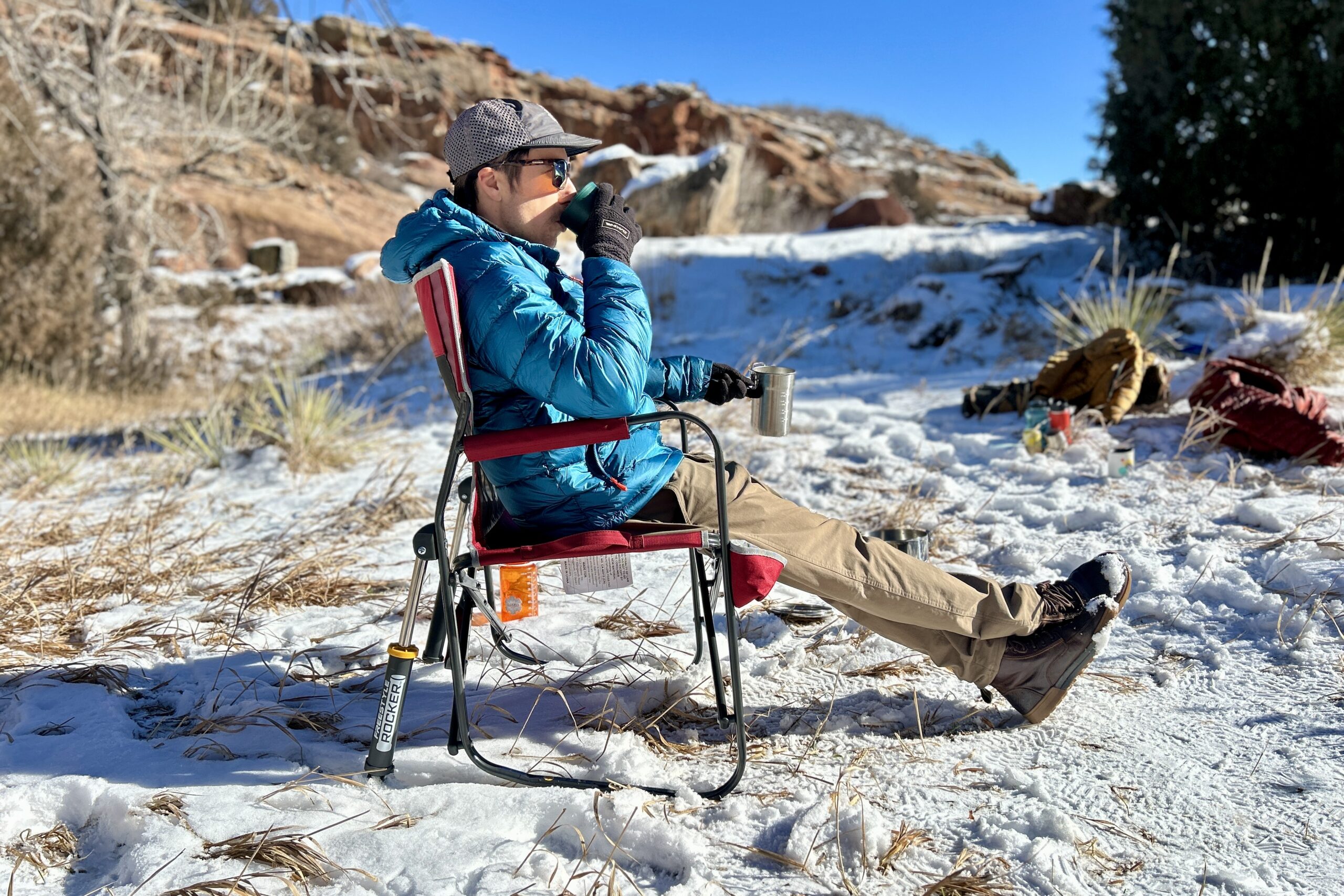 A side-profile image of man in a camping chair sipping on a mug with his legs crossed in front of him and snow on the ground.