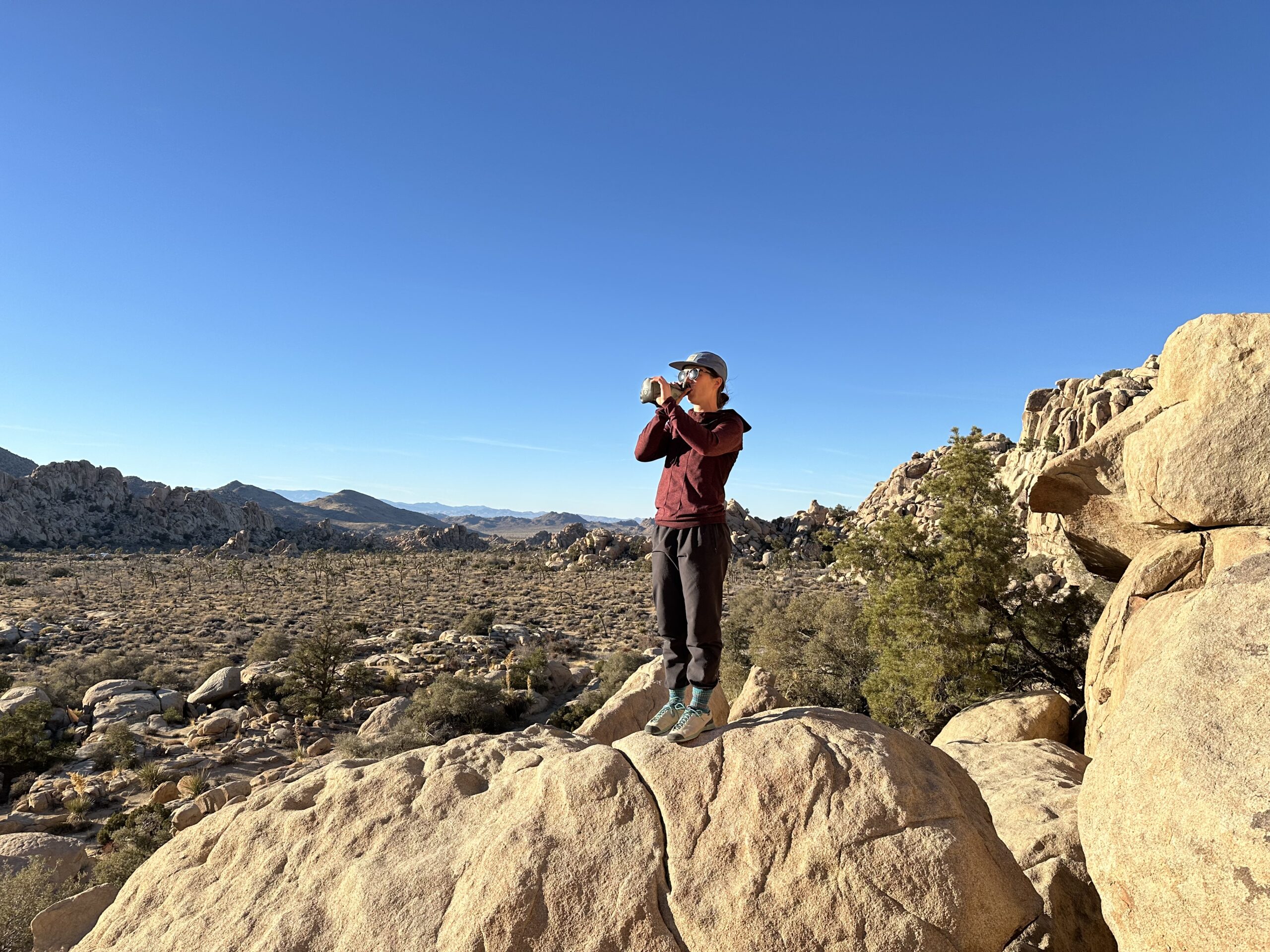 A far away shot of a hiker standing on a granite boulder while drinking out of the HydraPak Seeker water bag. The background shows the granite rock formations and scrub oak trees of Joshua Tree National Park.