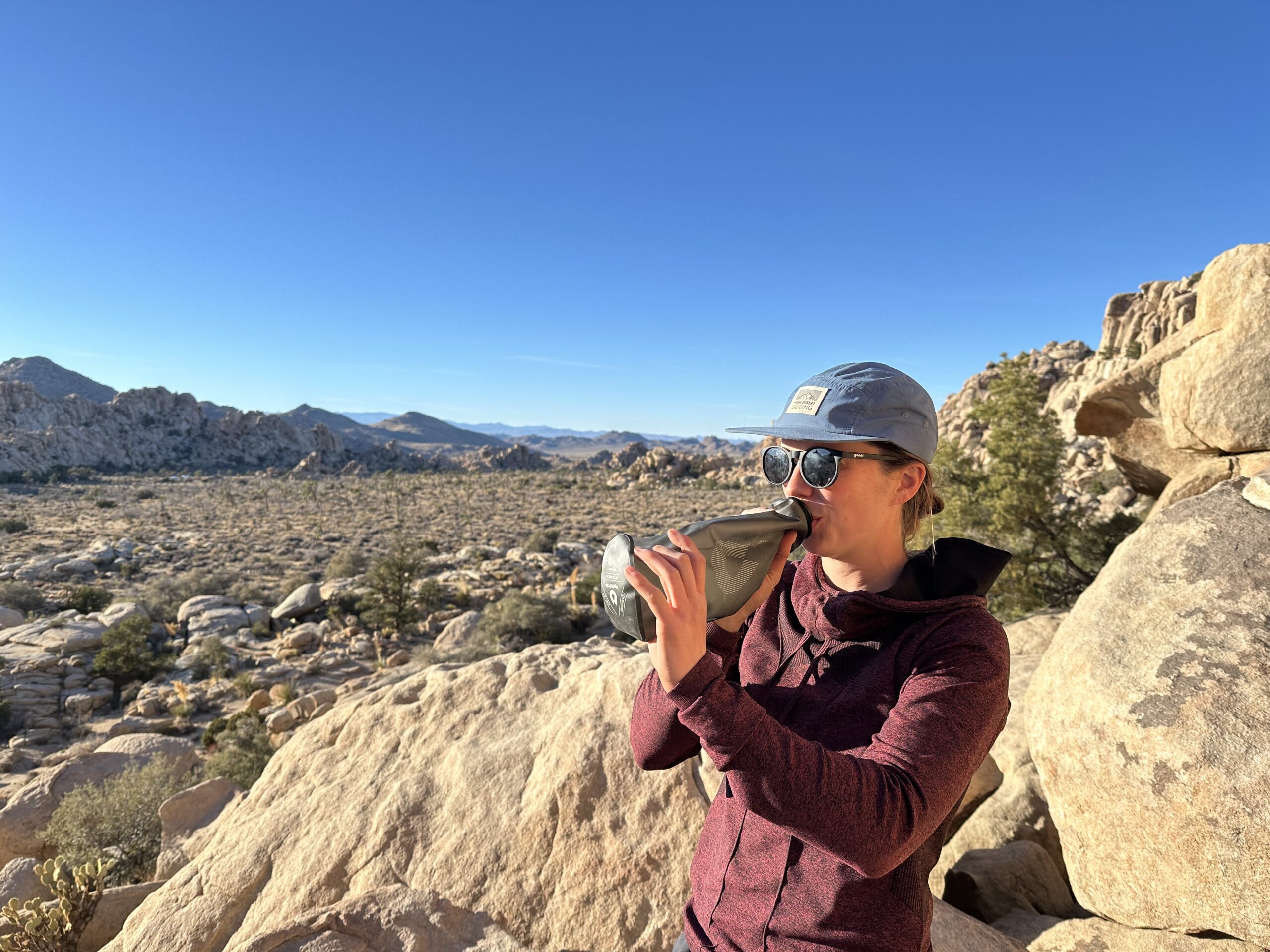 A person wearing sunglasses and a sun hat drinks from the HydraPak seeker water bag. The background is of Joshua Tree National Park, and it shows granite rock formations and some trees.