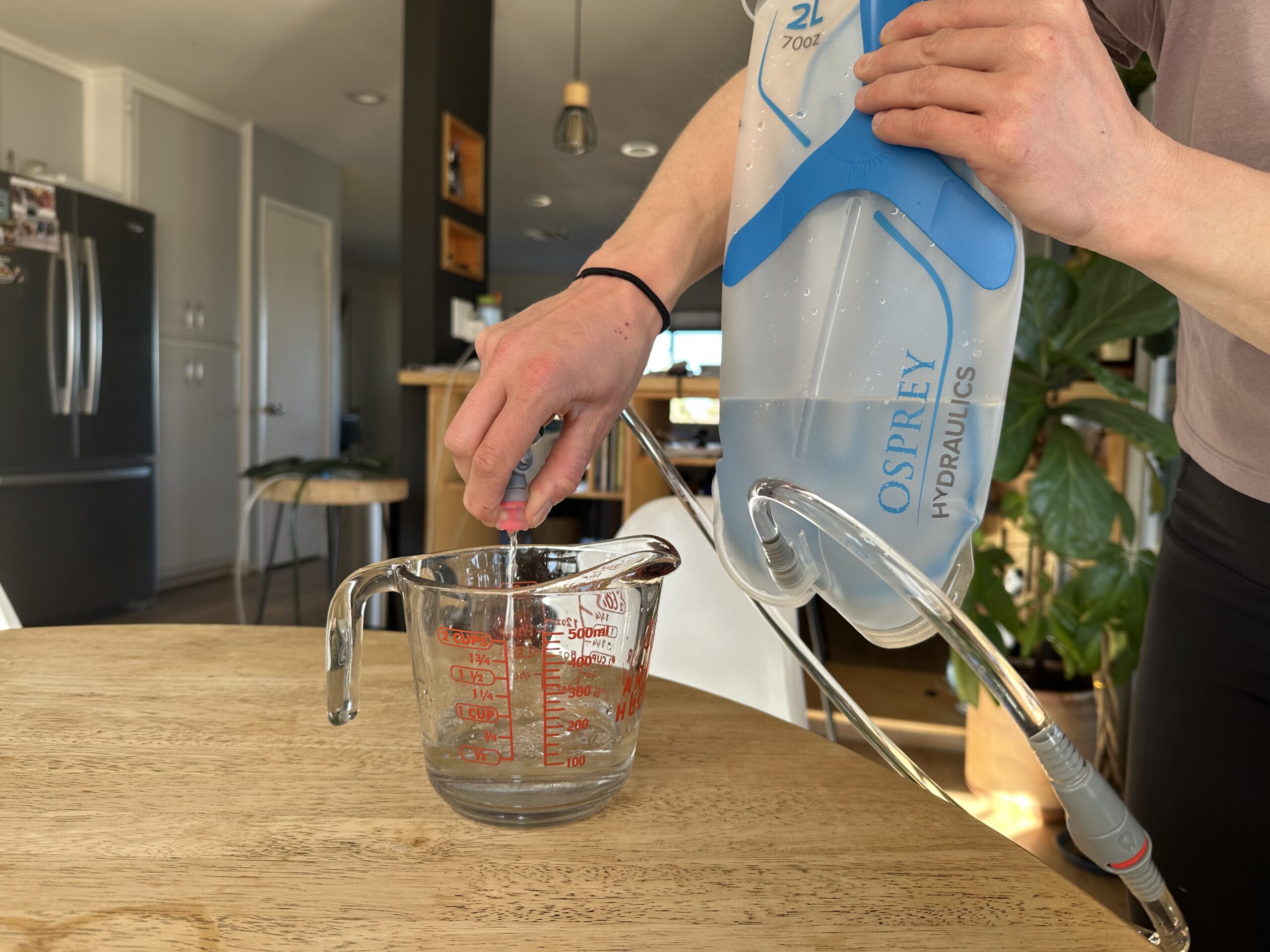 Close up photo of a person standing in a kitchen and holding the Osprey Hydraulics bladder in the air while squeezing the bite valve, allowing water to flow into a measuring cup.