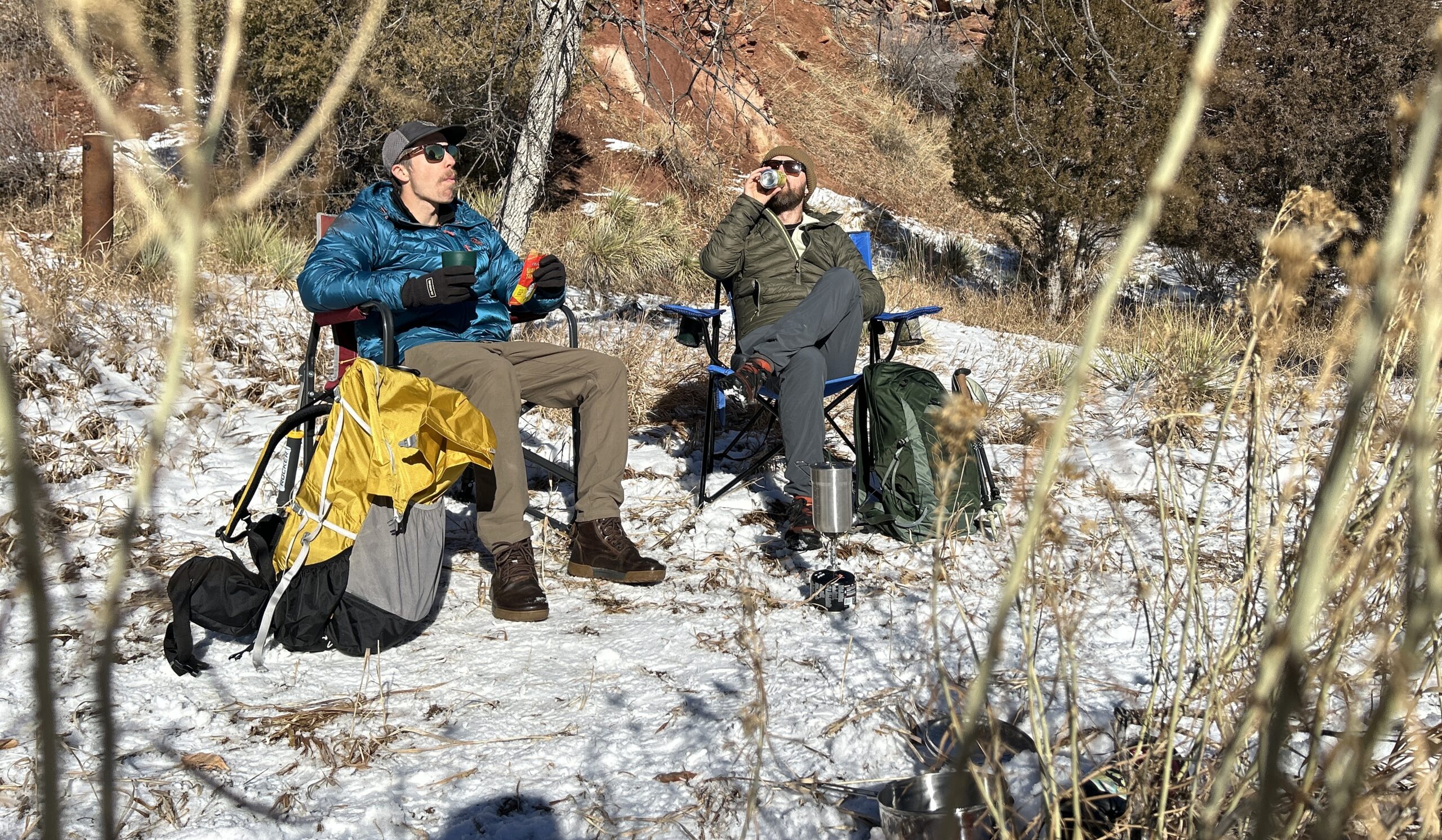 Two men sitting in camping chairs in a wintry desert landscape eat and drink and relax with their backpacks at their sides.