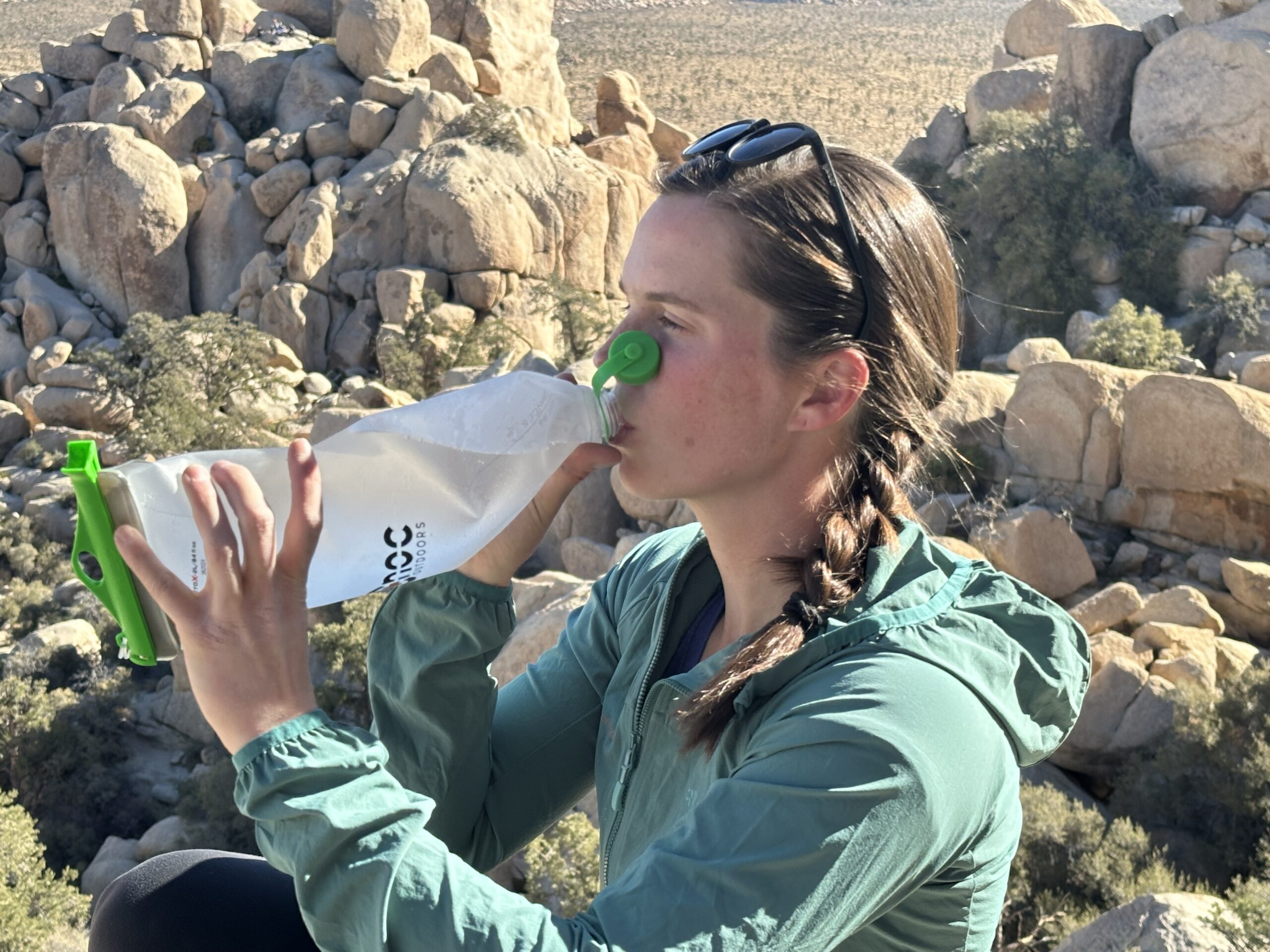 Close up of a hiker drinking from the CNOC VectoX water container, using two hands to hold it. The granite rock formations of Joshua Tree National Park are in the background.