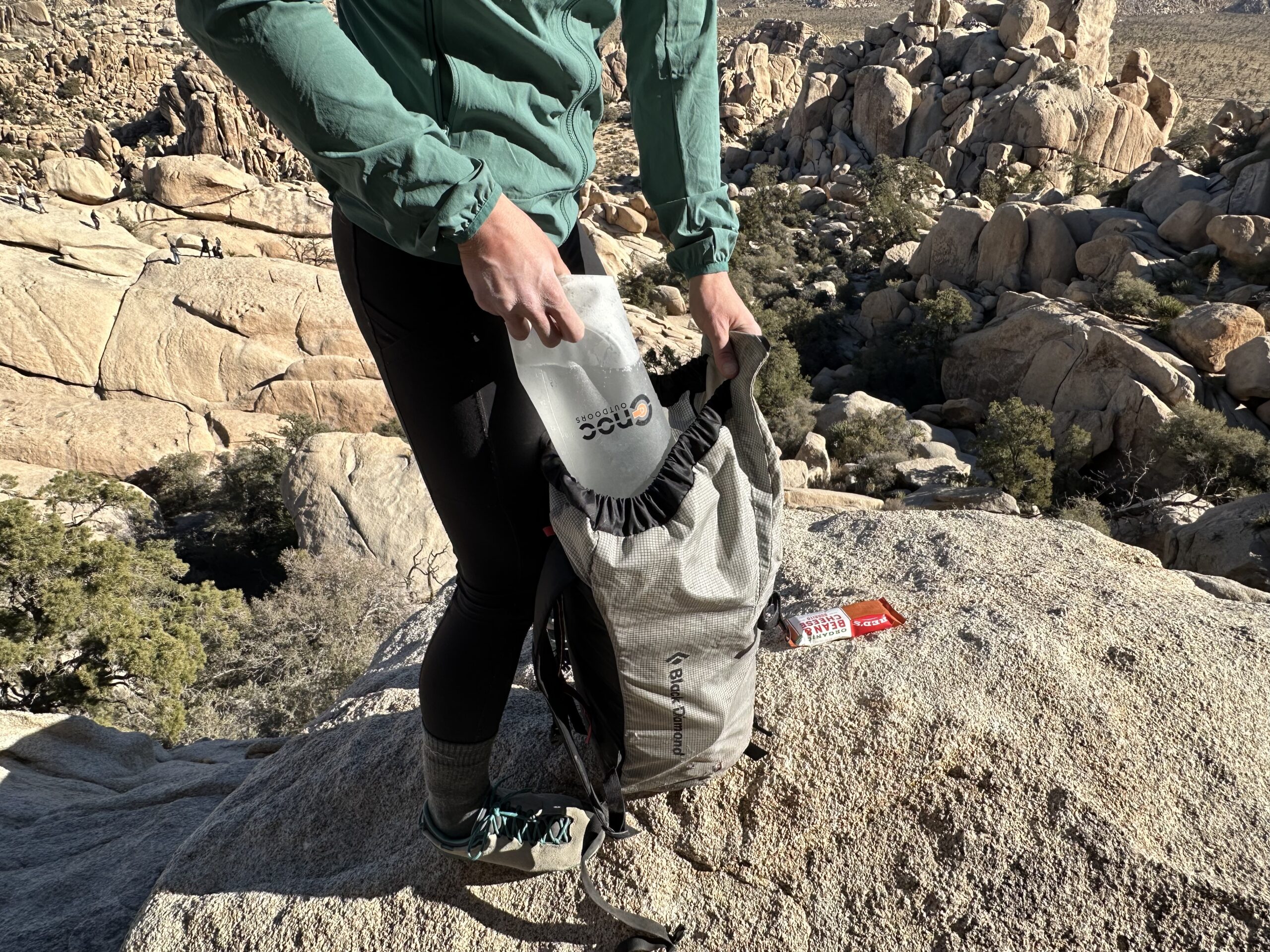 Close up of a hiker loading the CNOC VectoX into the top of her backpack. The background shows the granite rock formations of Joshua Tree National Park.