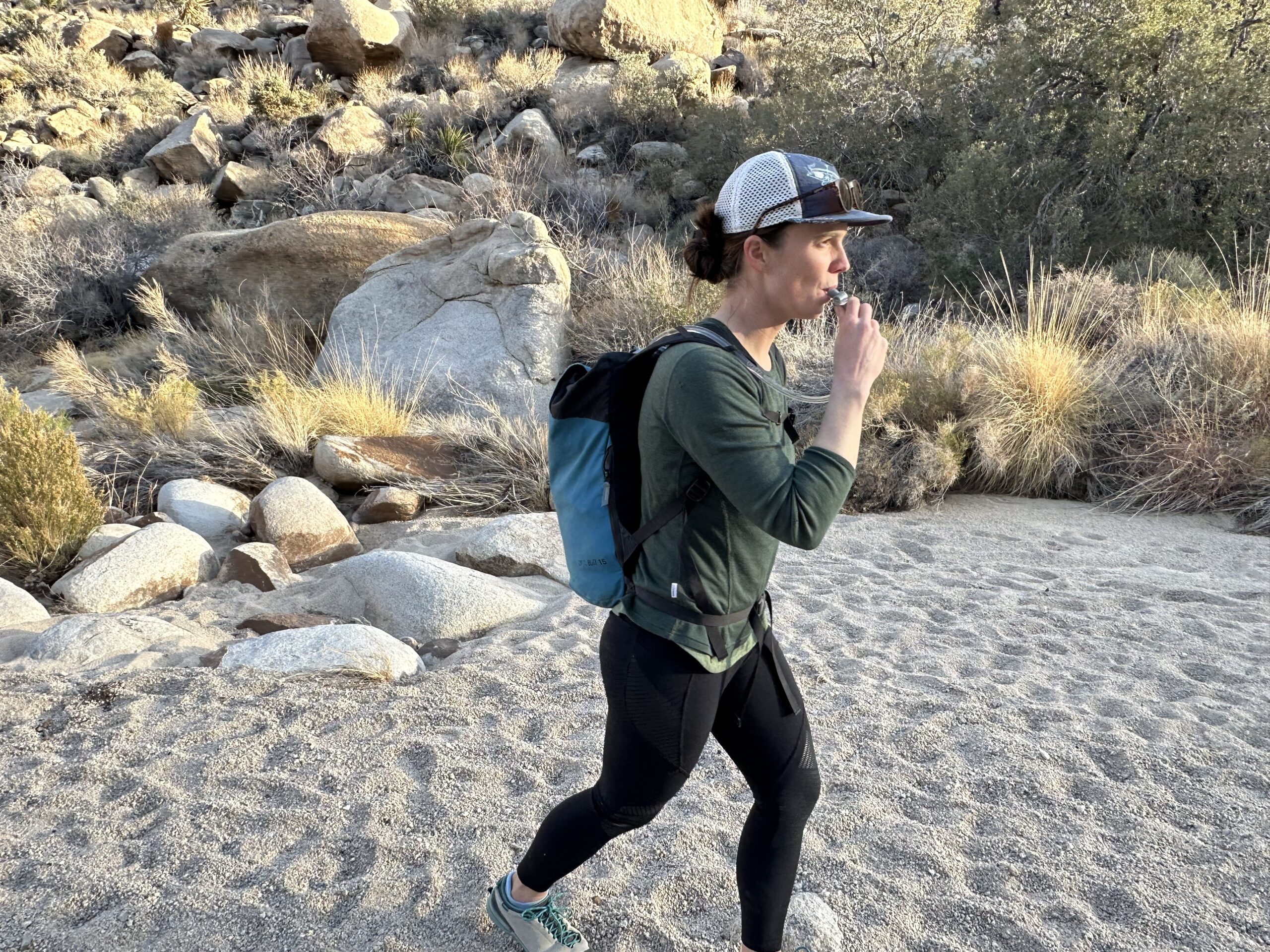 A hiker drinks from a hydration hose while walking through a sandy wash in Joshua Tree National Park