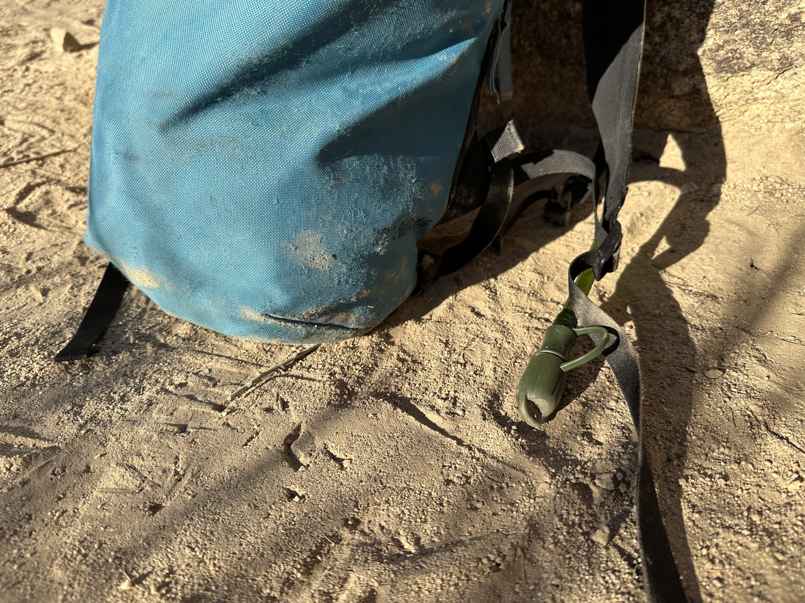 Close up of a hydration hose and bite valve coming out of a small backpack, all of which is sitting in the sand. The bite valve is protected with a cover.