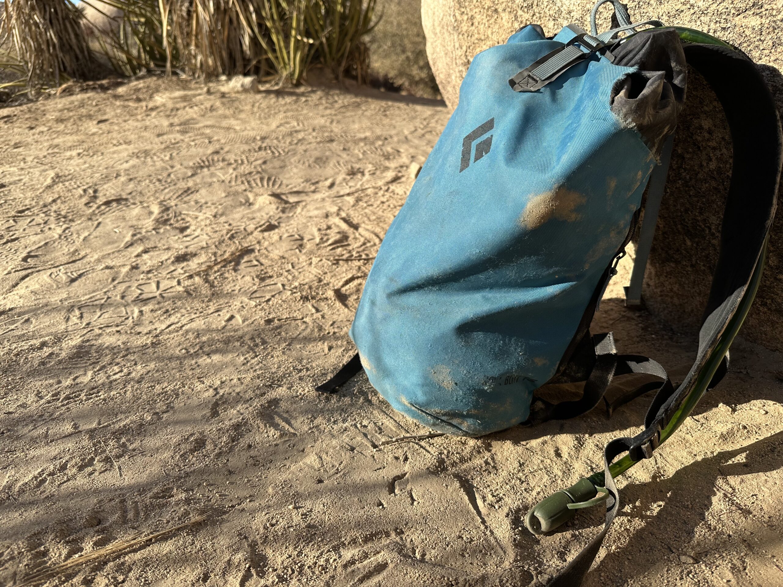 Close up photo of a bite valve and hose coming out of a small backpack. All are sitting in the sand, but the bite valve is protected with a cover