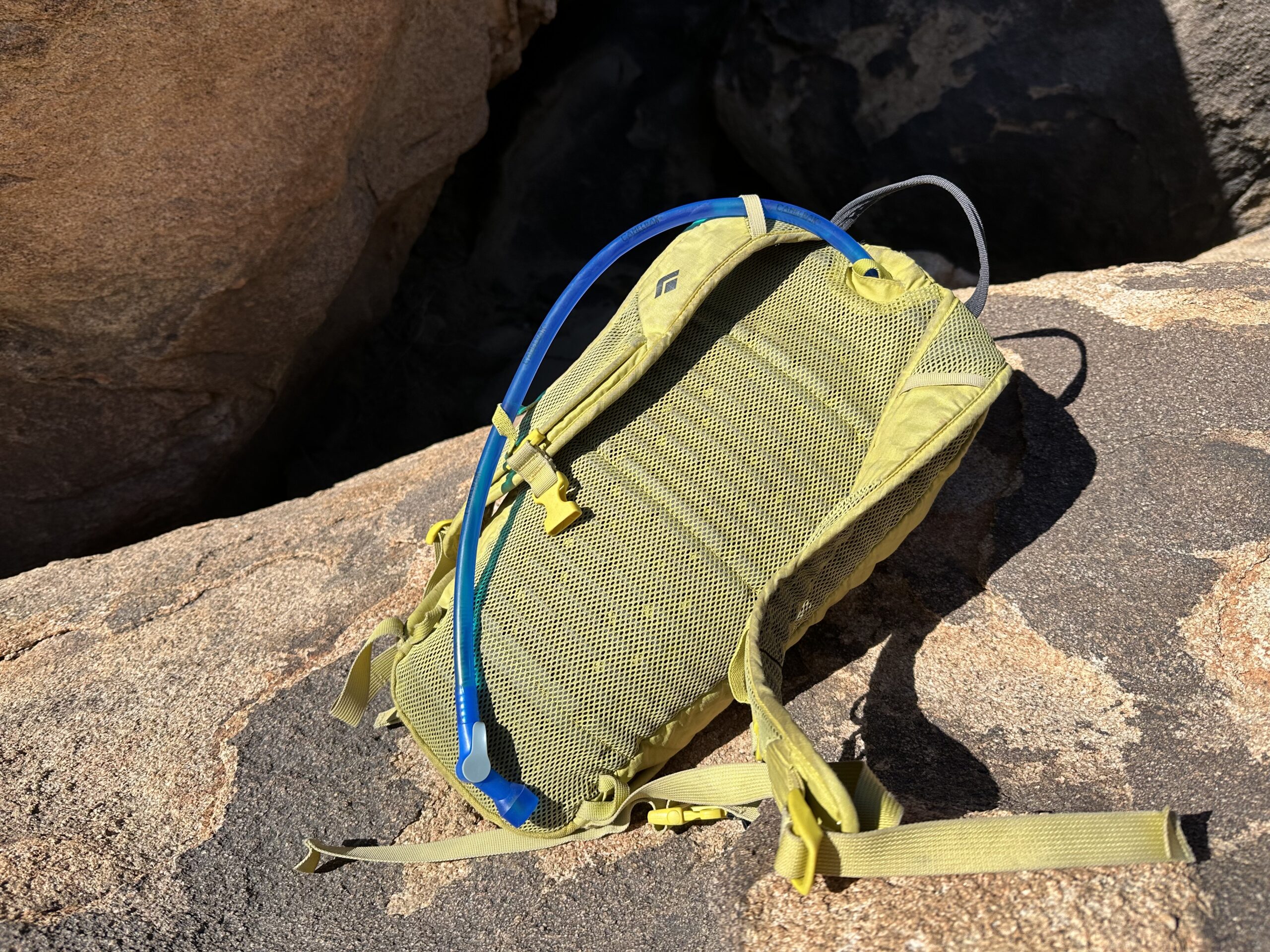 A close-up photo of the CamelBak Crux's hydration hose coming out of the back of a small yellow day pack. The pack is sitting on a large granite rock in the sun.