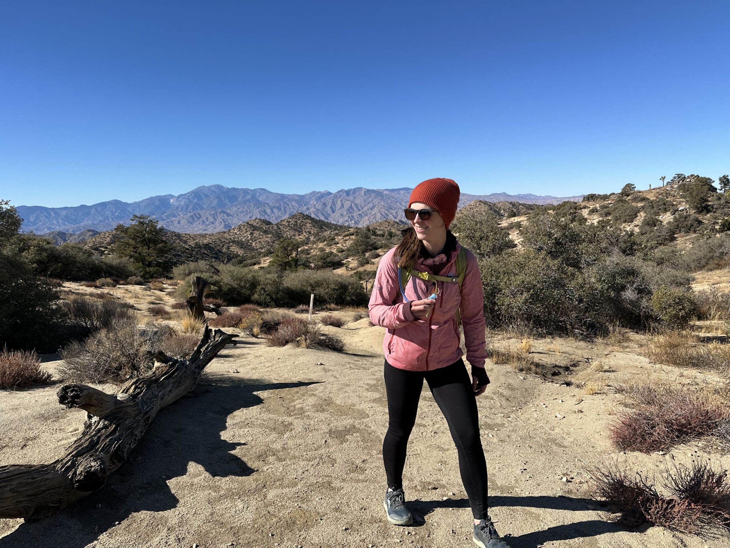 A hiker smiles while hiking on a trail in the Southern California desert. The trail is sandy with scrub oak bushes, and low, dry mountains are in the background.