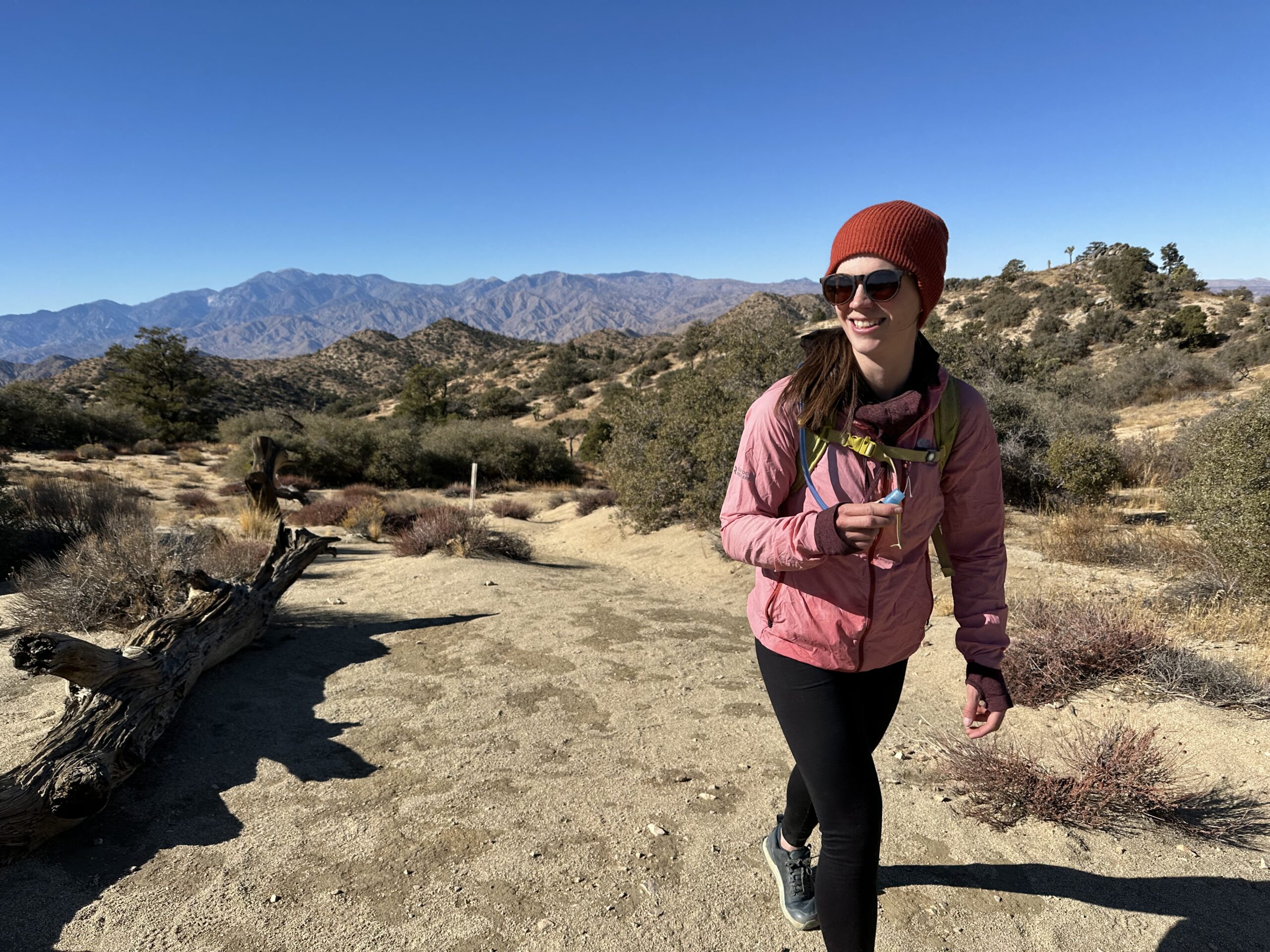 A hiker smiles while hiking on a trail in the Southern California desert. The trail is sandy with scrub oak bushes, and low, dry mountains are in the background.