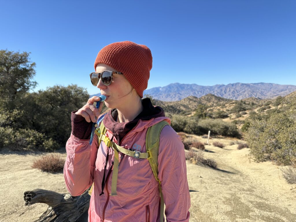 A hiker sips from a hydration hose on a hike in the Southern California Desert. Scrub oaks line the trail in the foreground, and brown mountains are in the background.