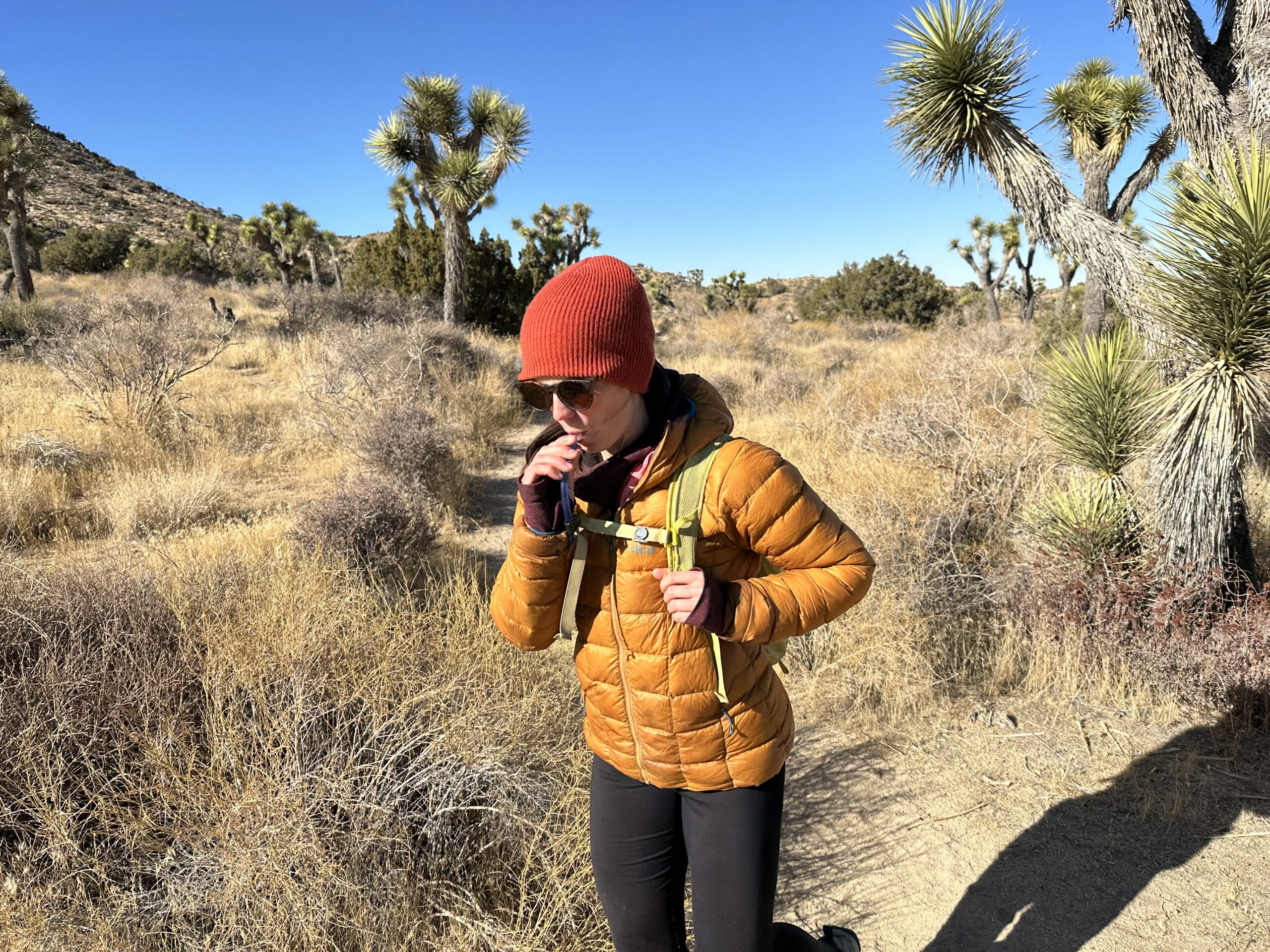 A hiker drinks from the Platypus Hoser while hiking on a trail in the desert with Joshua Trees in the background.